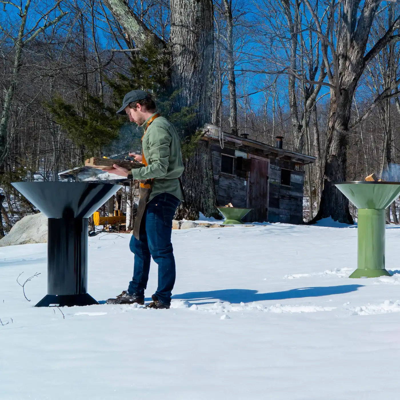 founder putting wood on black Vuuris Onyx next to a green Vuuris Sage with a snowy background and a shed in the background