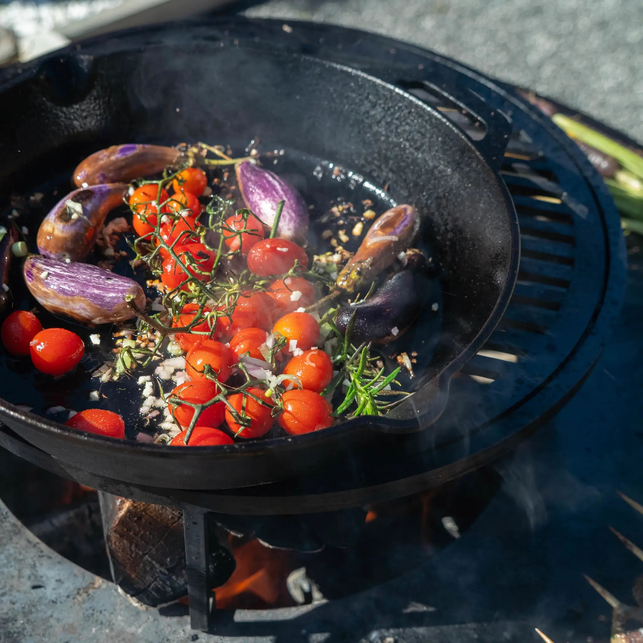 Daylight photo of a cast iron pan on top of the Vuuris grill placed on a Vuuris cooking plate. Tomatoes, eggplant and herbs are in the pan with a fire underneath.