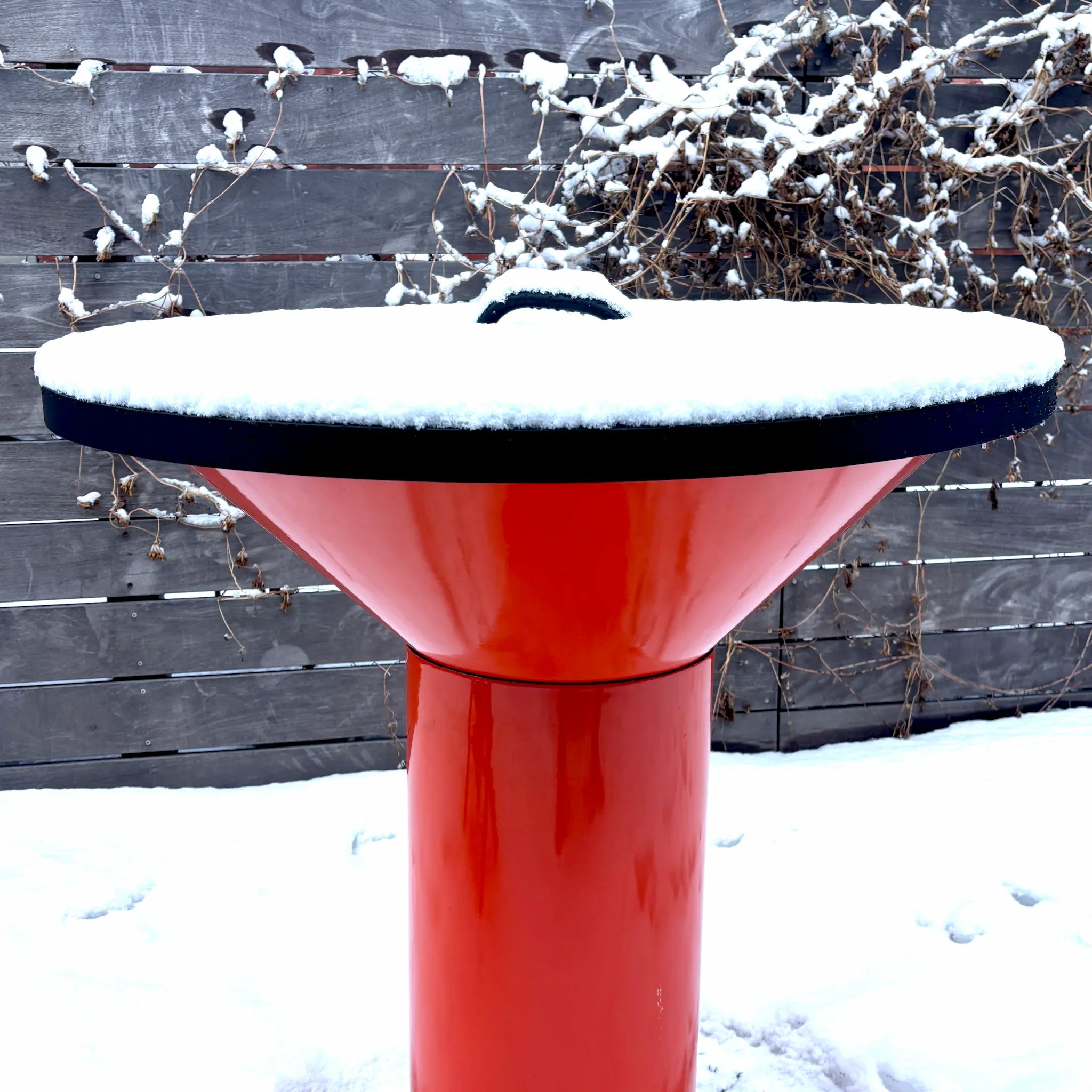 Vuuris matte black steel lid fitted on the orange Vuuris Tiger against a wooden fence background with snow on the ground and a layer of snow on the lid, illustrating its use case