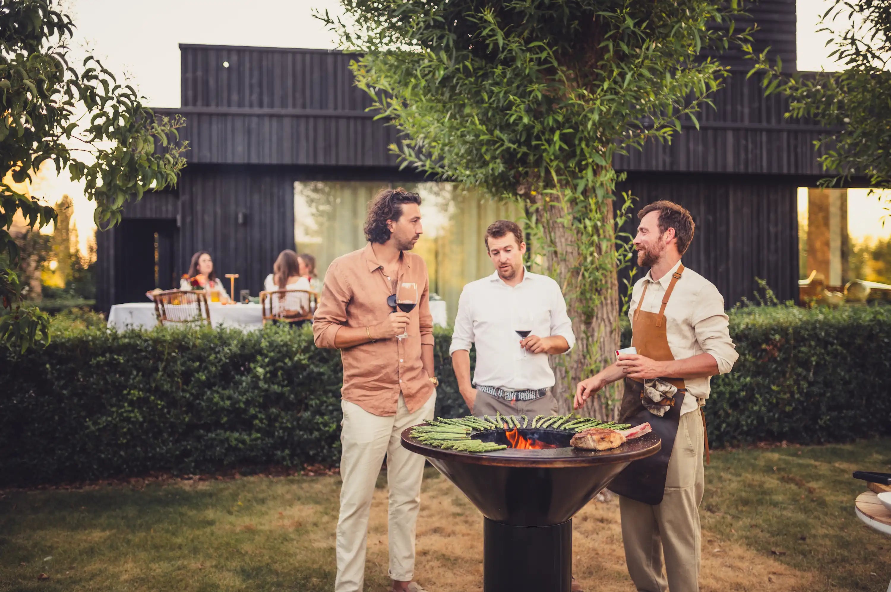 Three men standing outdoors at a black Vuuris Onyx barbecue grill in a backyard setting, with a modern dark wooden house and trees in the background.