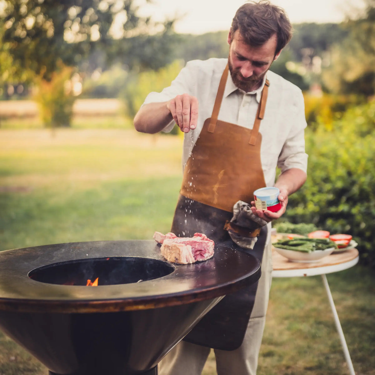 A man wearing a brown leather Vuuris apron over a white shirt grills a steak outdoors on a round black barbecue grill Onyx Vuuris, with a small table holding vegetables in the background.