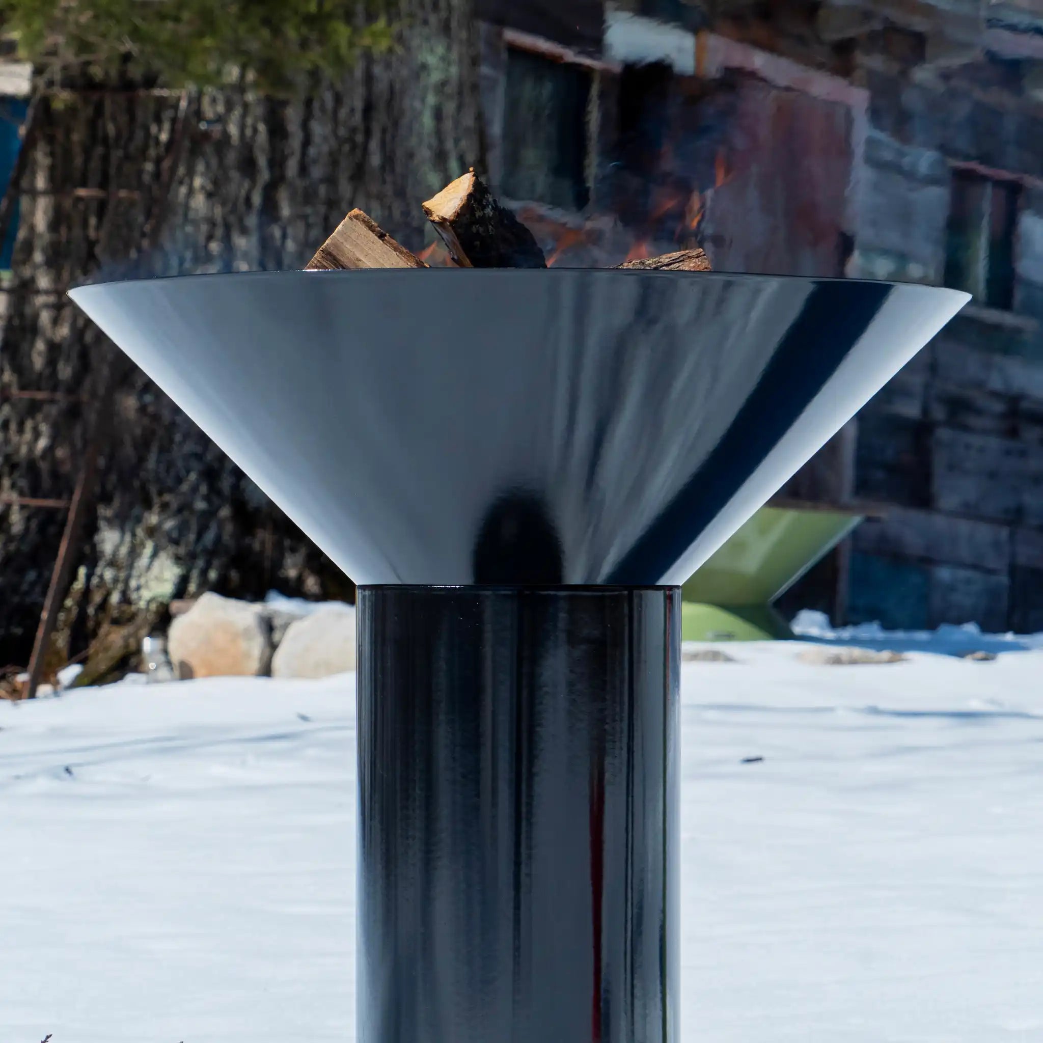 close up of Vuuris Onyx outdoor fire pit in deep black porcelain enamel mounted on the large base with a fire inside against a background of a wooden shed in the snow