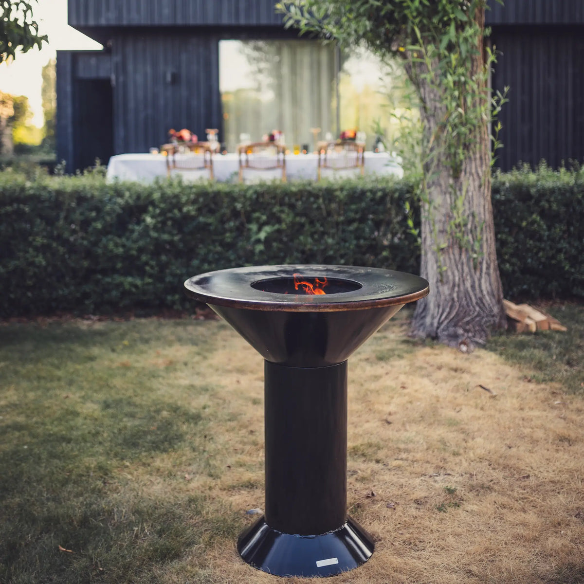 A black metal pedestal fire pit Vuuris Onyx with a visible flame, situated outdoors on a lawn in front of a hedge and a dark wooden house.