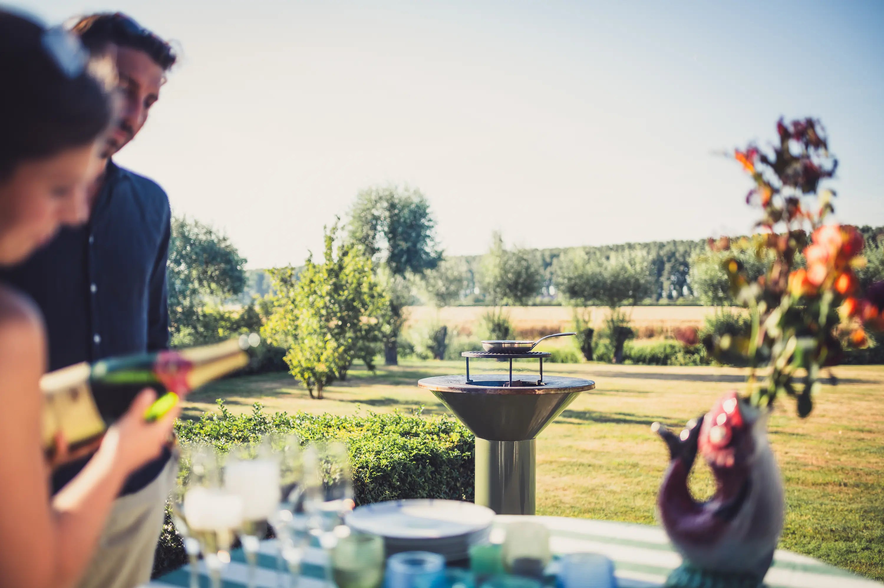 An outdoor garden party scene with a man and a woman in the foreground, one holding a champagne bottle, with a table, glasses, and a decorative figure in the foreground and a grassy lawn, trees, and a blue sky in the background with a Vuuris Sage green barbecue fire pit in the center.