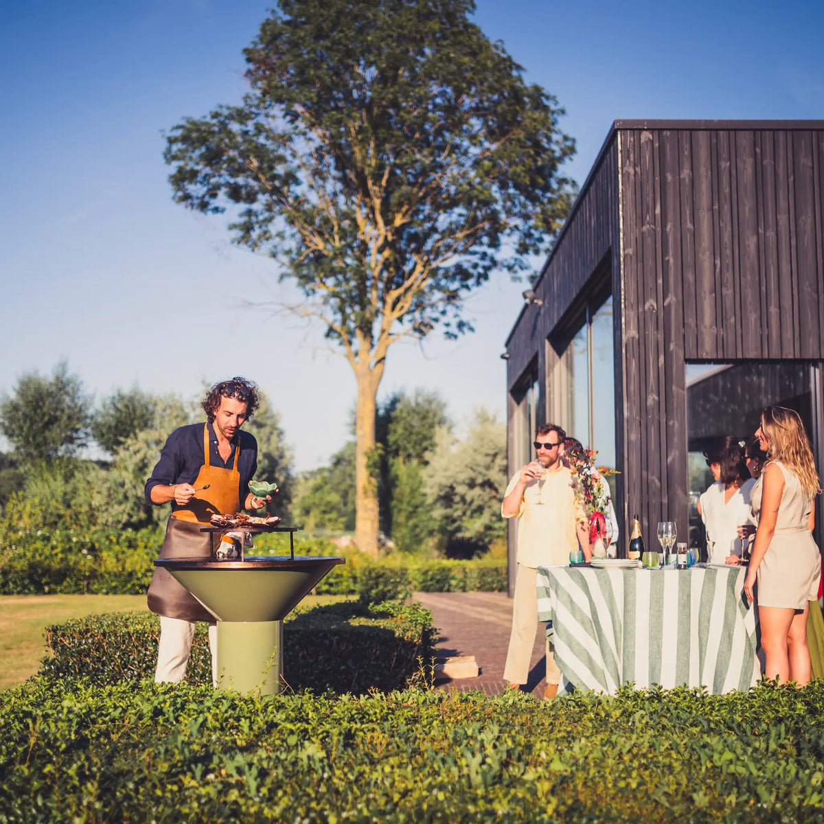 Outdoor wedding or event scene with a man grilling on a green Sage Vuuris bbq in the foreground, while guests stand by a striped tablecloth and dark wood building under a blue sky.