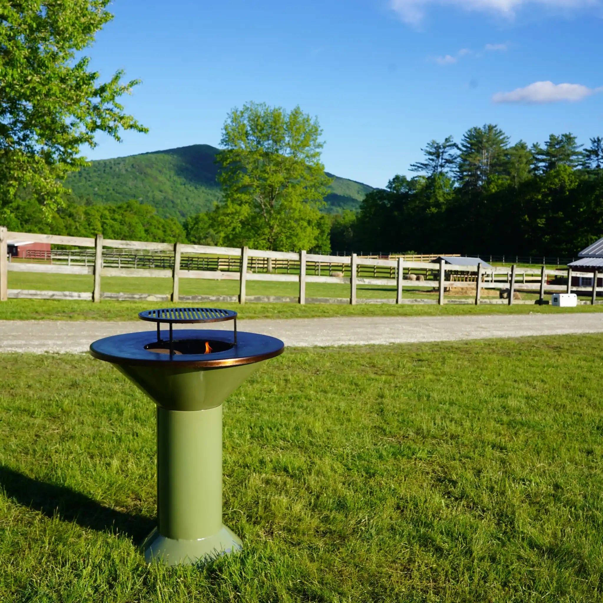 Vuuris Sage on large base setup with fire inside and grill on top of cooking plate against a background of a farm with green grass, mountain in background and some fencing