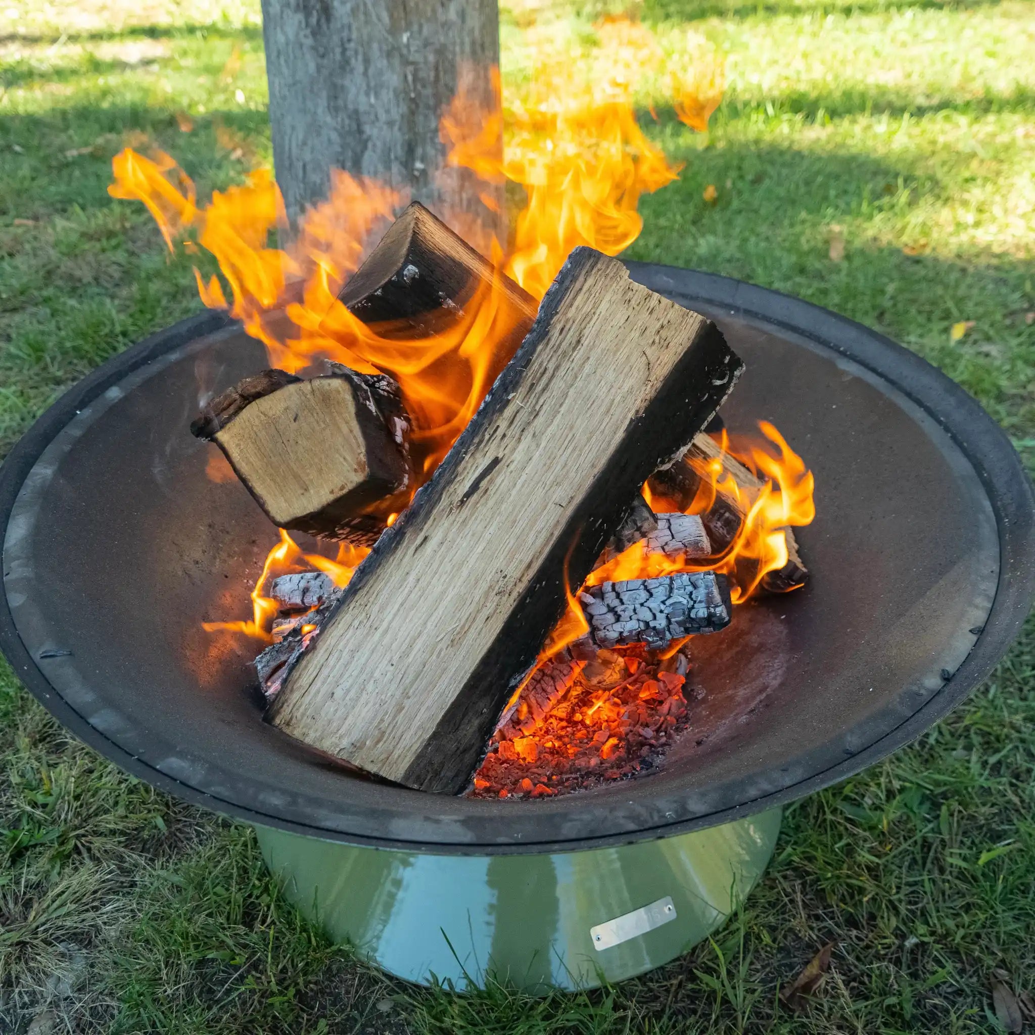 close up of Vuuris Sage outdoor fire pit in earthy green porcelain enamel mounted on small base at lounge height with a fire inside, standing on grass in a garden