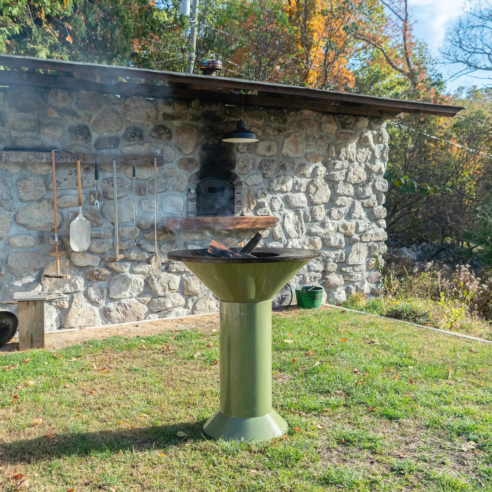 Vuuris Sage outdoor fire pit in earthy green porcelain enamel mounted on large base at counter height with a cooking plate on top and a fire inside against a background of a stone house standing on grass of the garden