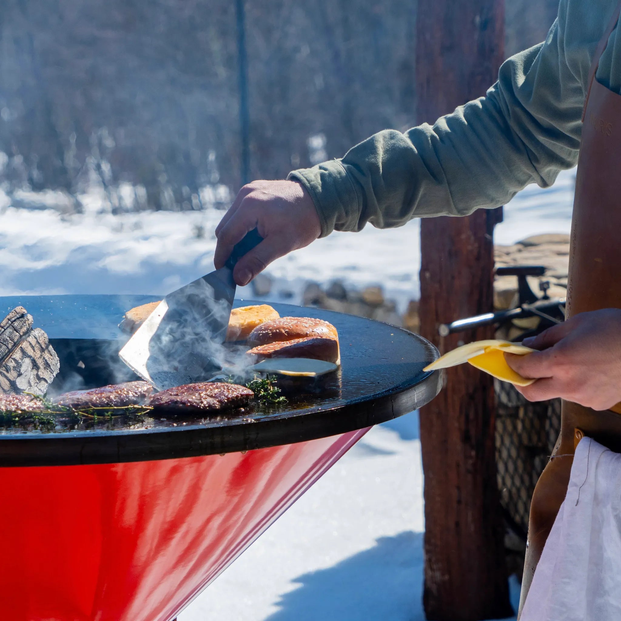 Close-up detail of the Vuuris long-blade burger turner, in action on the orange Vuuris Tiger flipping a burger that is cooking against a snowy garden background