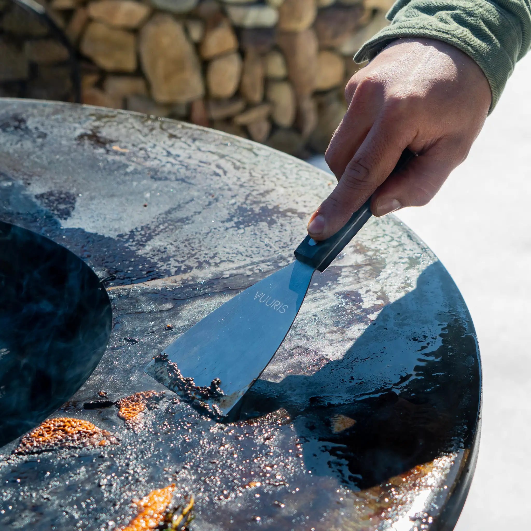 Vuuris straight scraper spatula in action scraping food remains off the cooking plate, with a clear view of the engraved Vuuris logo and themoplastic handle