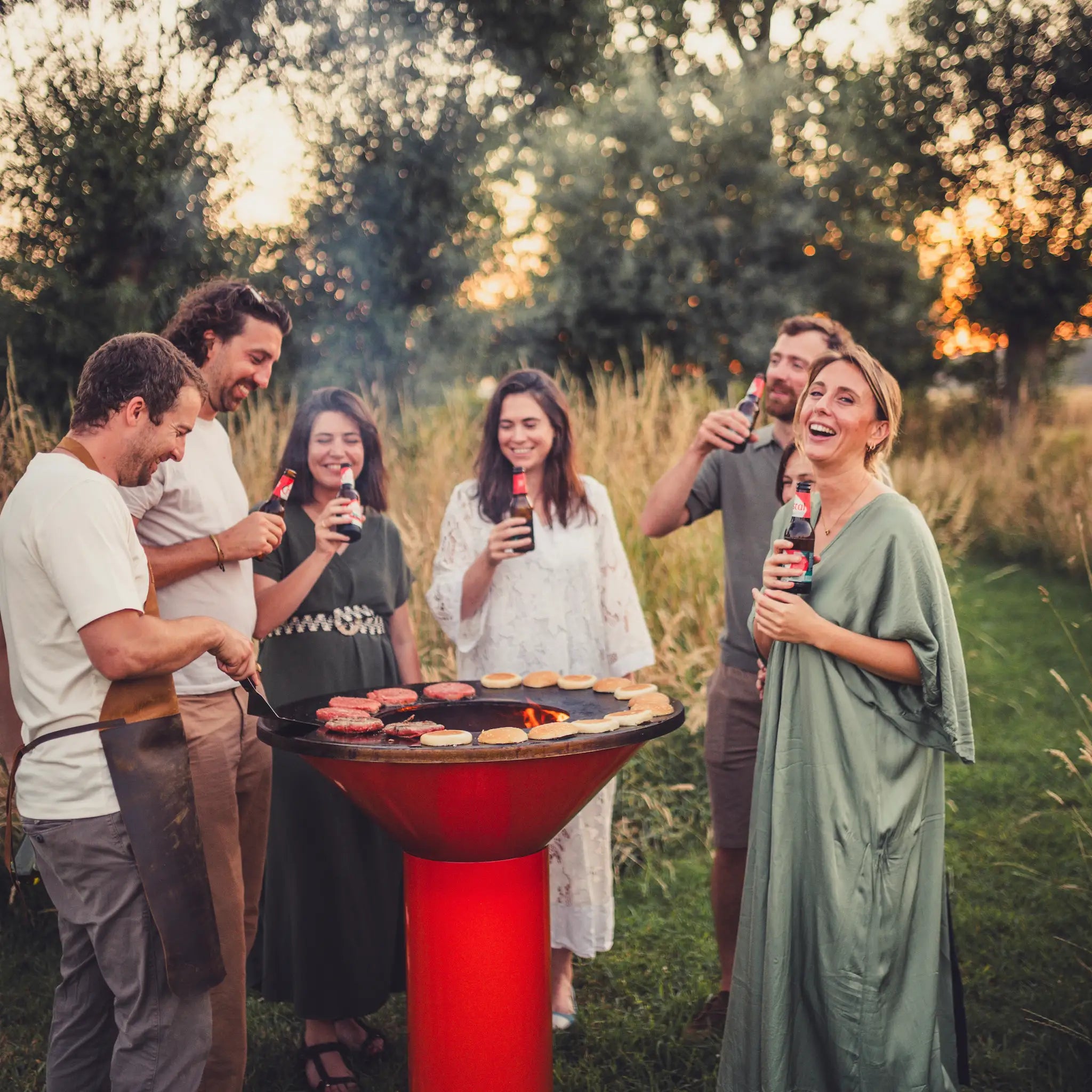 A group of five people gathered outdoors at sunset around an orange Vuuris Tiger freestanding barbecue grill, holding drinks and grilling burgers. The scene is set in a grassy backyard with trees in the background.