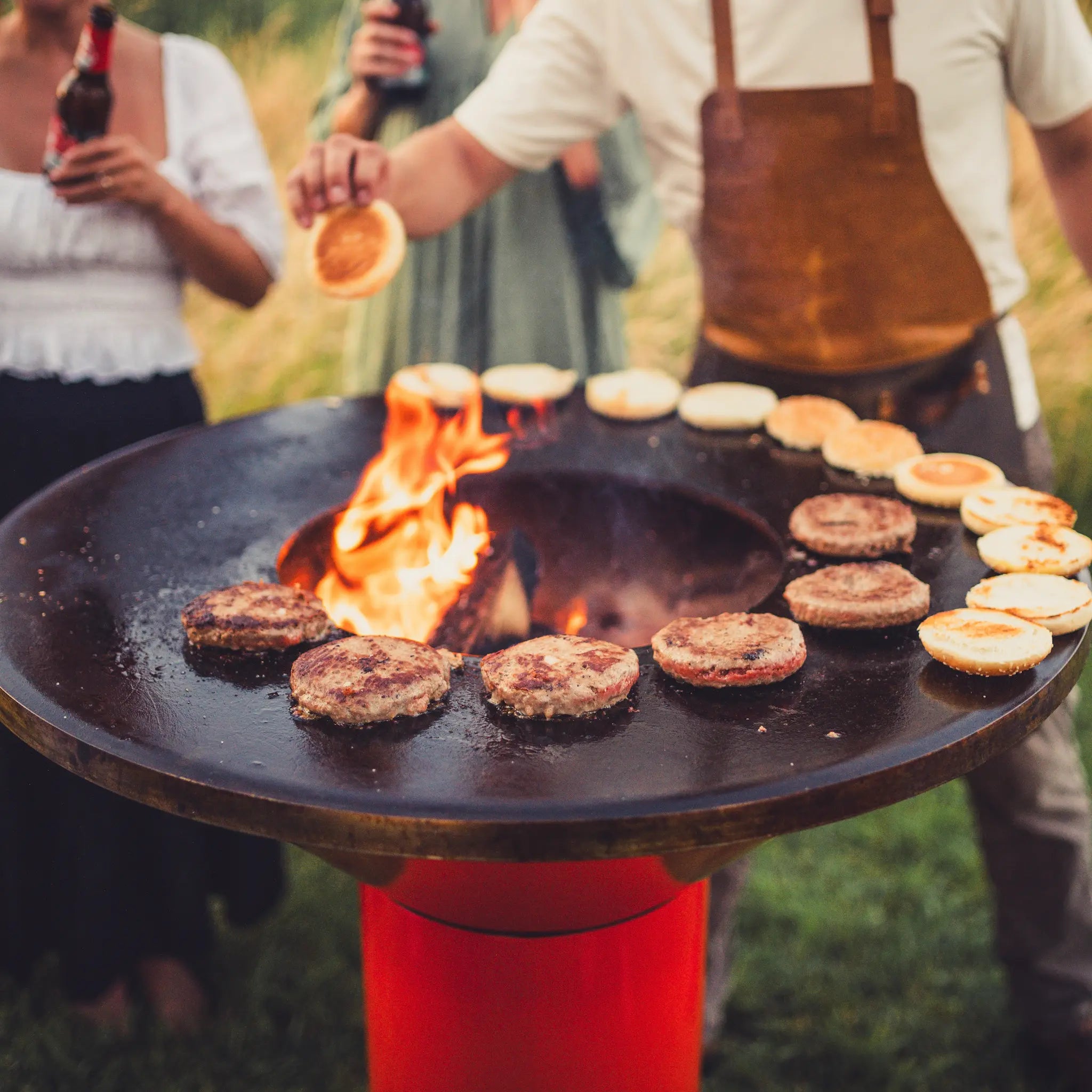 An orange, round Vuuris Tiger outdoor cooking grill with a black top surface and orange base, featuring burger patties and buns cooking over an open flame, with people standing around it in a grassy outdoor setting.