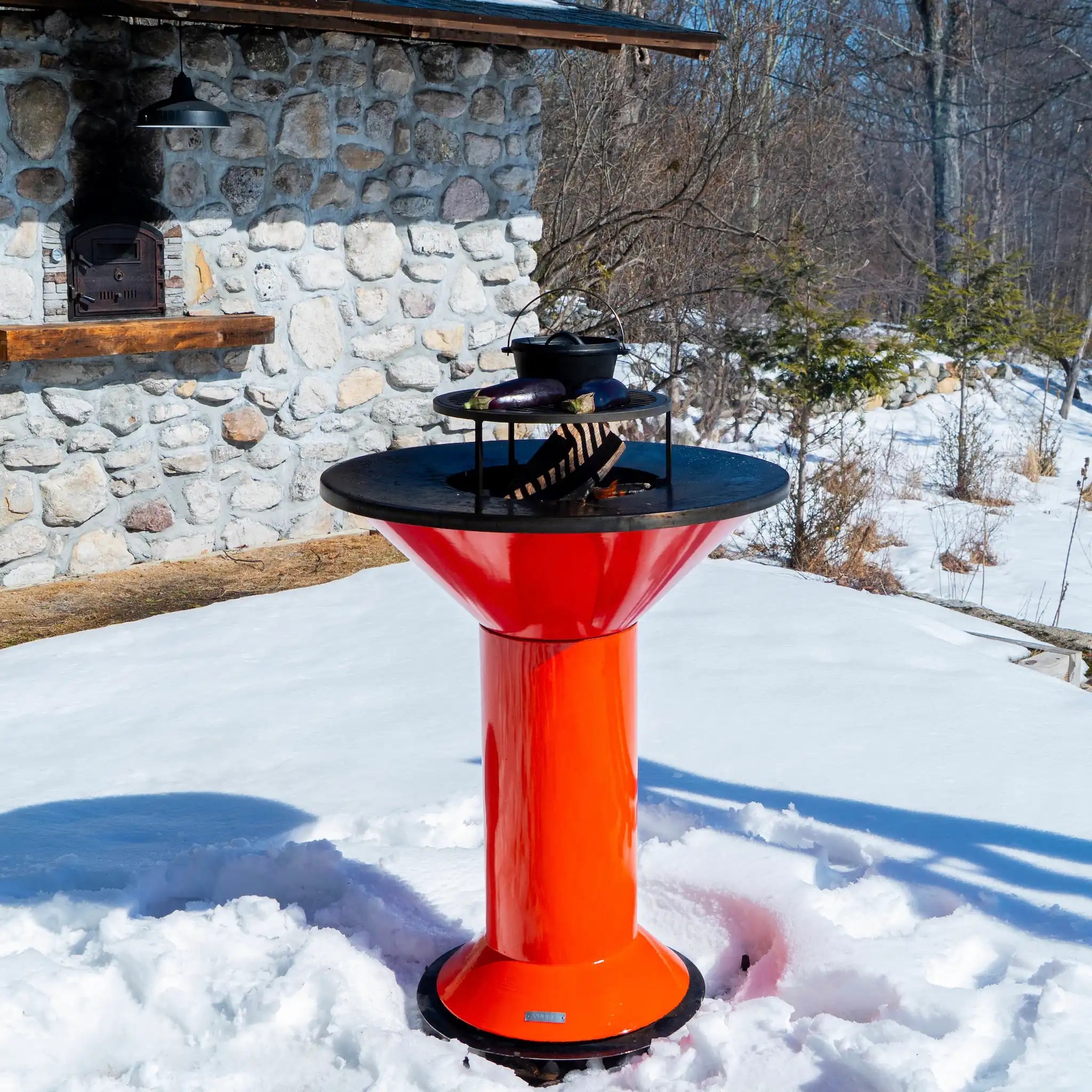 Vuuris Tiger outdoor fire pit in vibrant orange porcelain enamel with a grill on top and a fire inside - against a background of a stone house with snow on ground