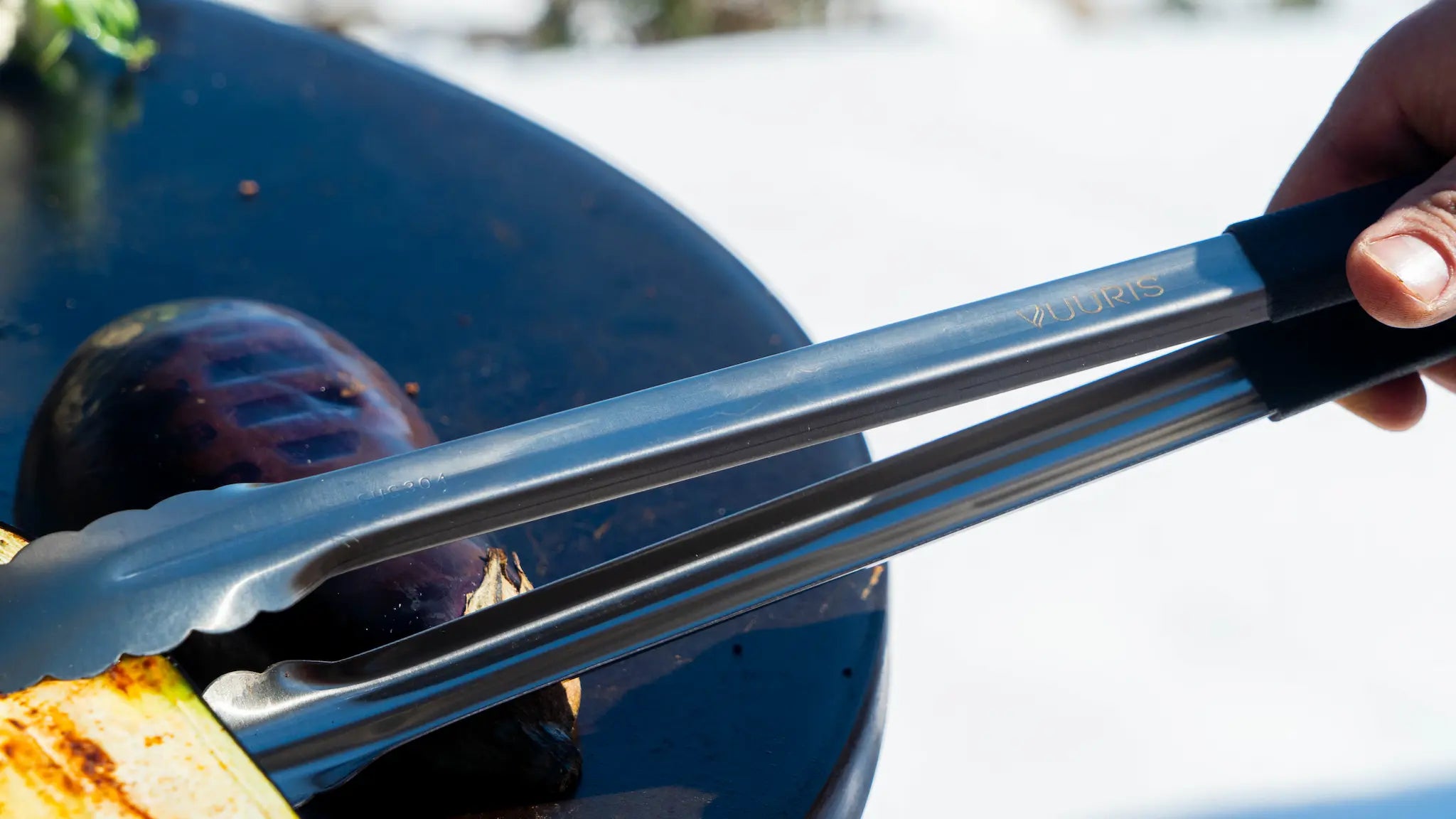 Vuuris 16-inch stainless steel tongs, detail shot showing the ergonomic matte black thermoplastic grip and laser-engraved Vuuris logo while flipping an aubergine on the Vuuris cooking plate