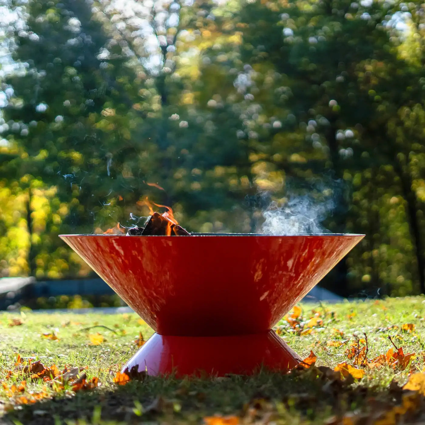 Orange Tiger Vuuris fire pit with flames and smoke in a grassy area with trees in the background