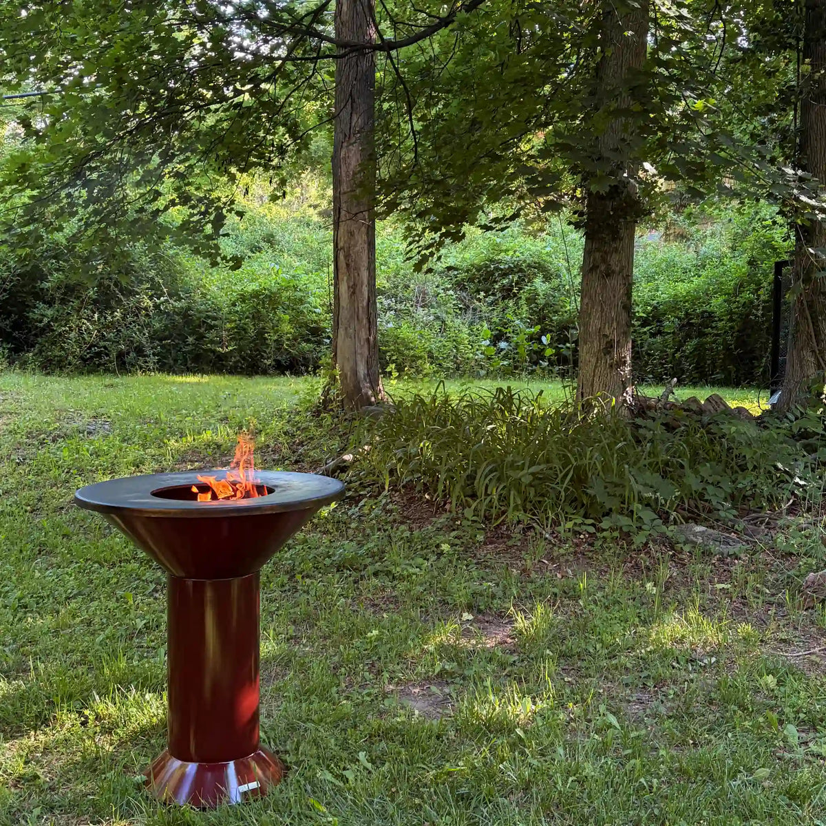 Garnet bordeaux Vuuris bbq with flames in it against a background of grass and trees.