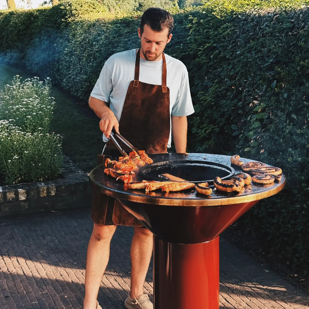 A man wearing a brown leather apron grills meat on a round outdoor fire pit Vuuris Garnet grill in a garden setting.