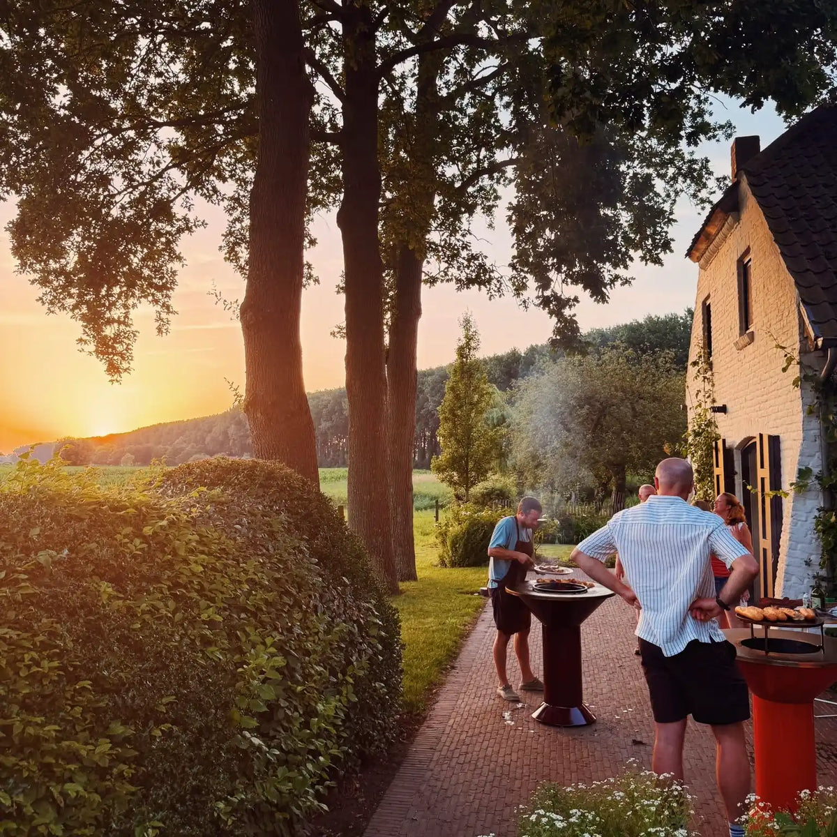 Man cookiing meat on the Vuuris Garnet bbq at sunset next to a white house and field and trees