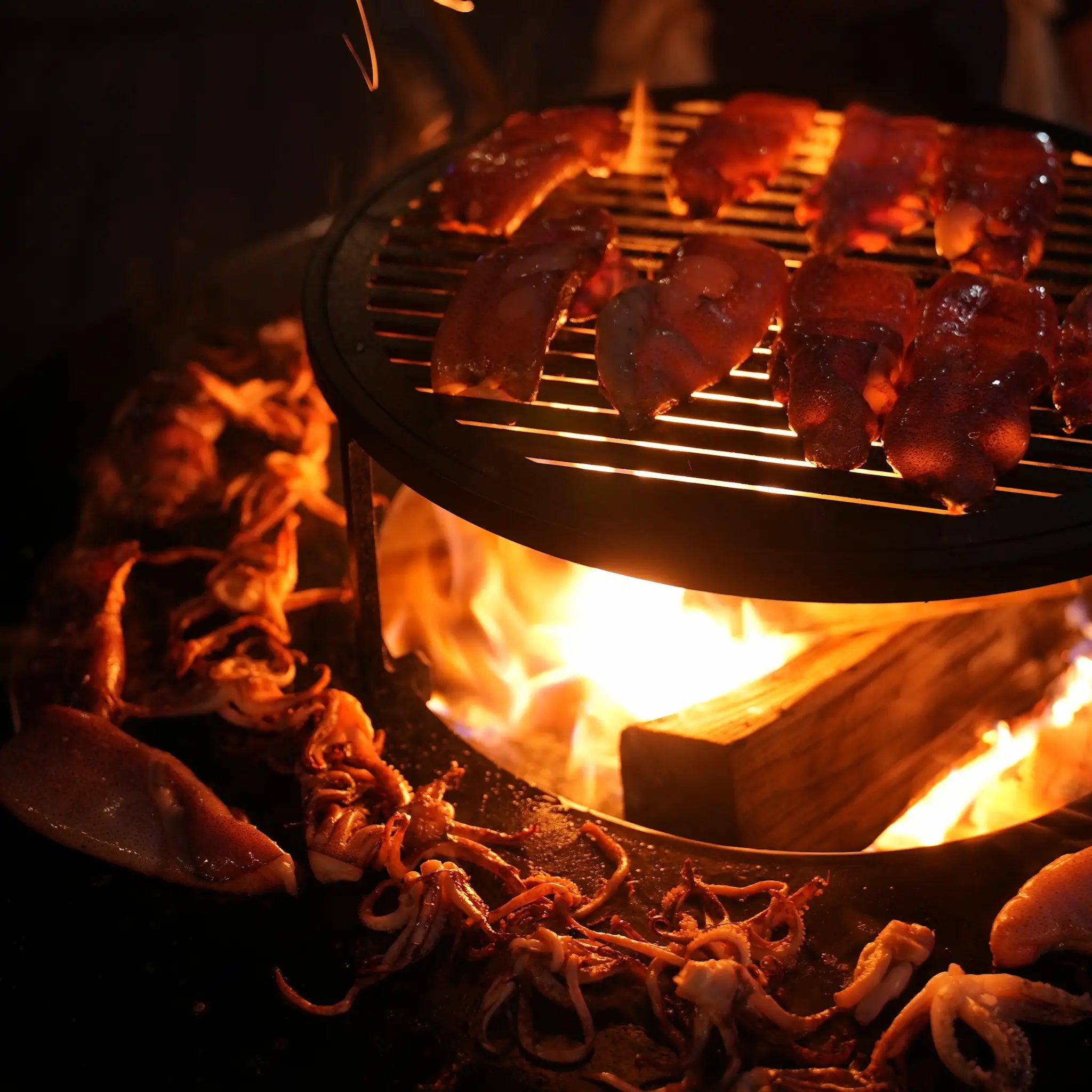 Close up of squid being grilled on the Vuuris grill and cooking plate with a burning fire in the middle in darkness