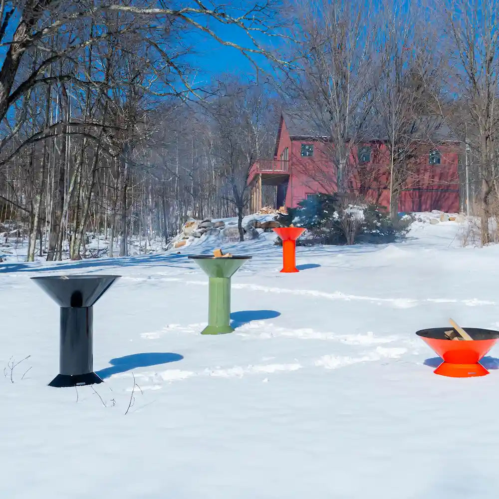 A wide shot of a field full of snow with a red house in the background with 4 Vuuris products that are on fire, a orange Tiger, green Sage and black Onyx in the large base setup and one orange Tiger in small base setup