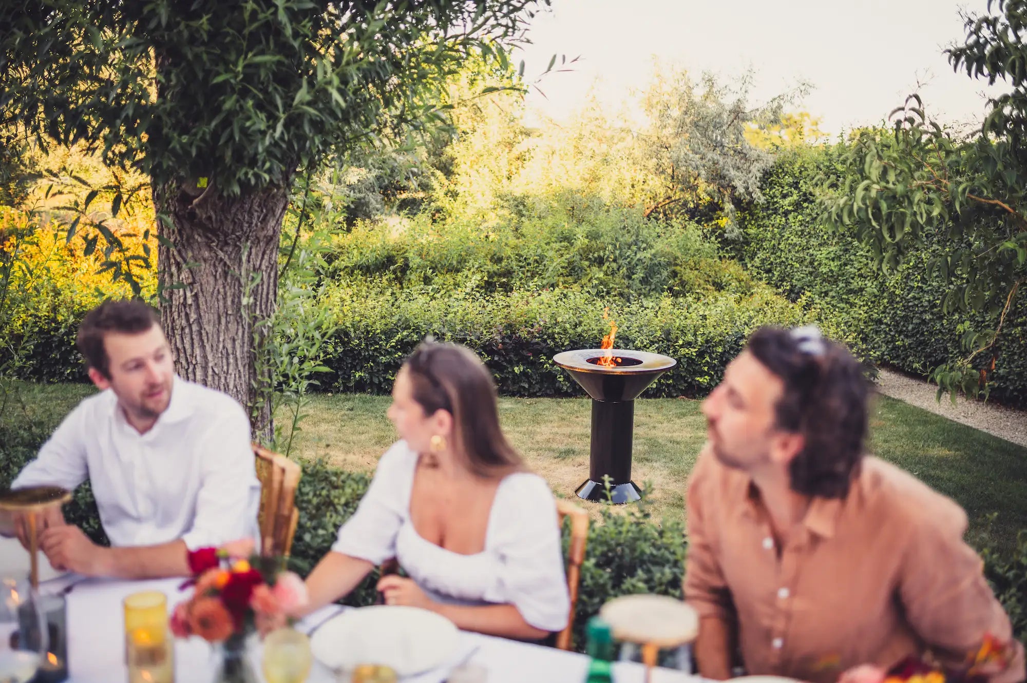 Three people sitting at an outdoor dining table in a garden setting, with trees, greenery, and a Vuuris Onyx large base fire pit bbq in the background.