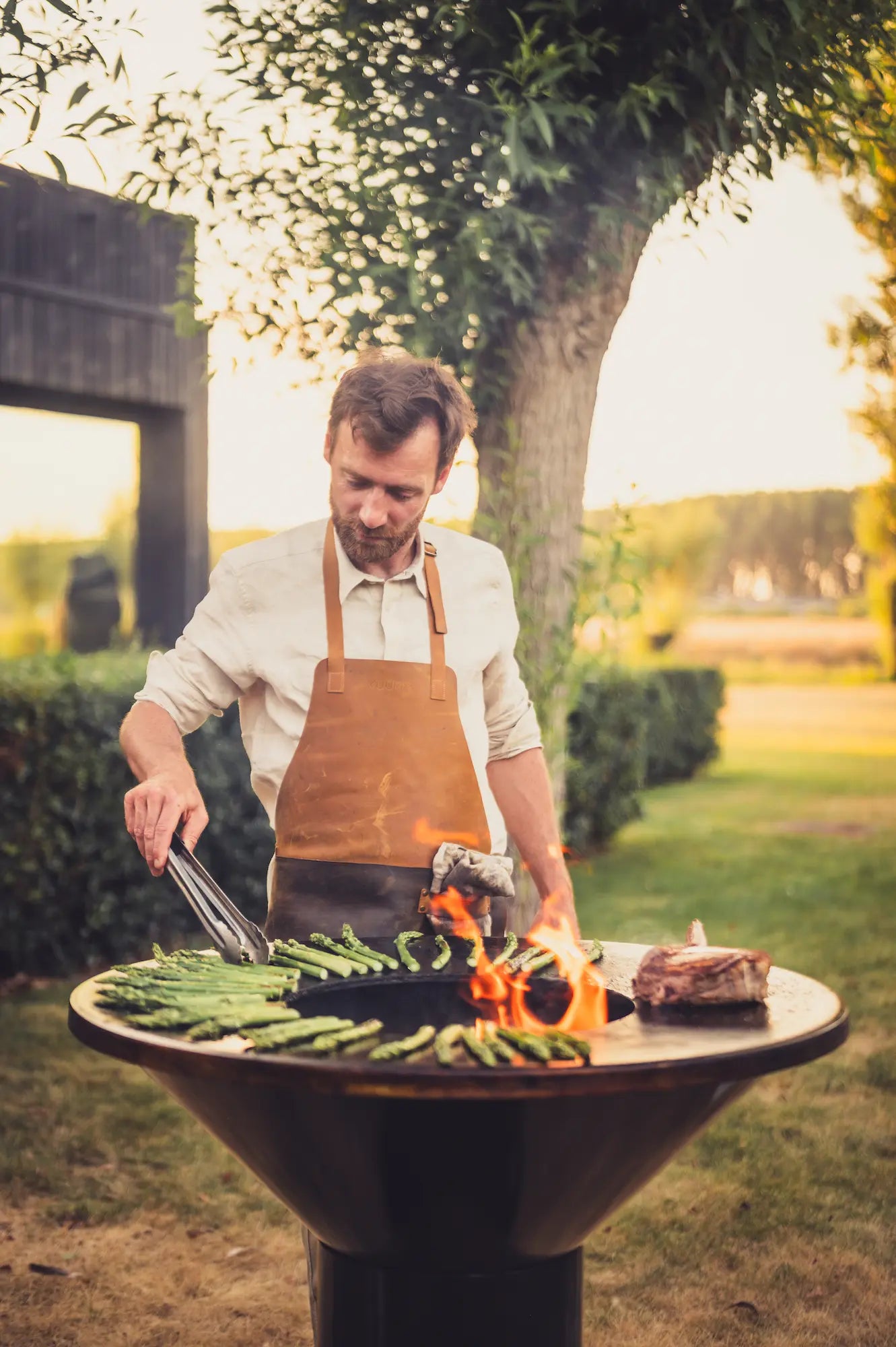 A man wearing a white shirt and brown leather Vuuris apron grills food on a round outdoor Vuuris Onyx barbecue in a park setting with trees and grass.