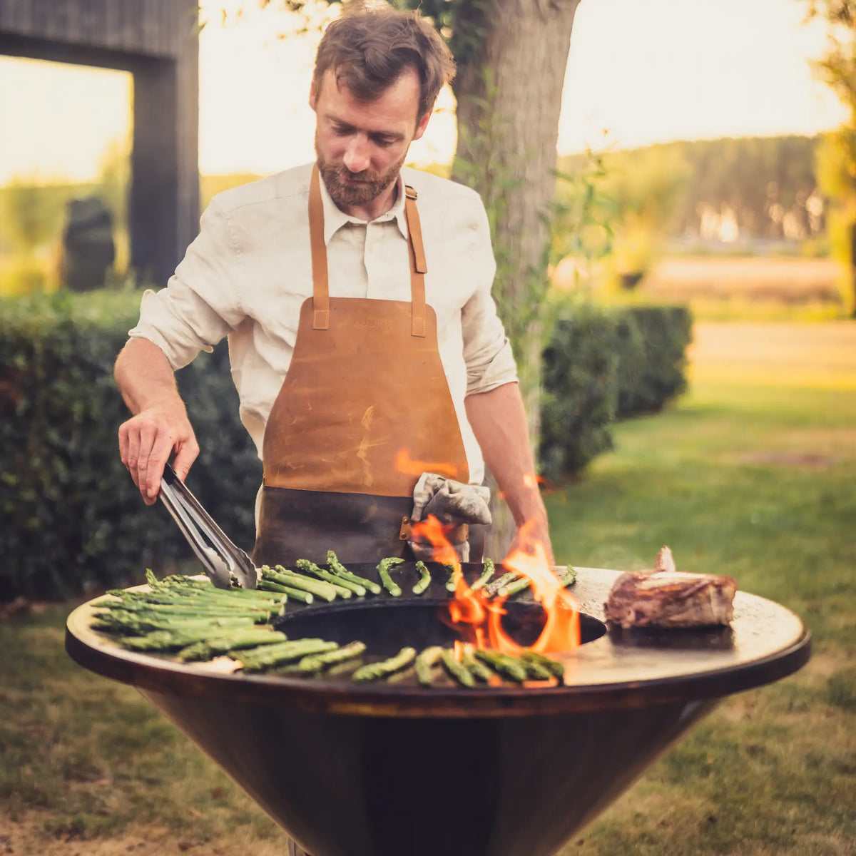 A man wearing a brown leather apron grills food on an outdoor round fire pit Onyx Vuuris with green asparagus and a steak, set in a grassy backyard.