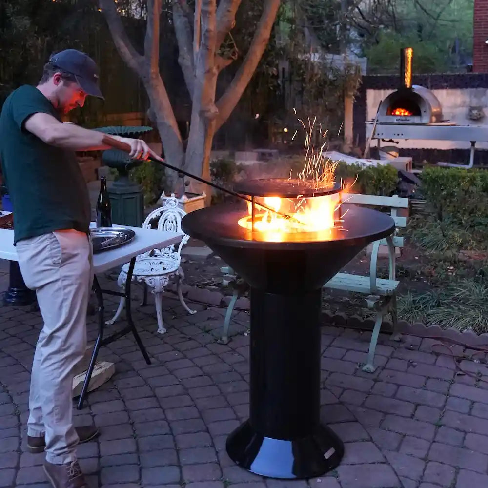 Person using a black blow poke with wooden handle to stir a burning fire in the Vuuris Onyx on the large base in a garden setting.