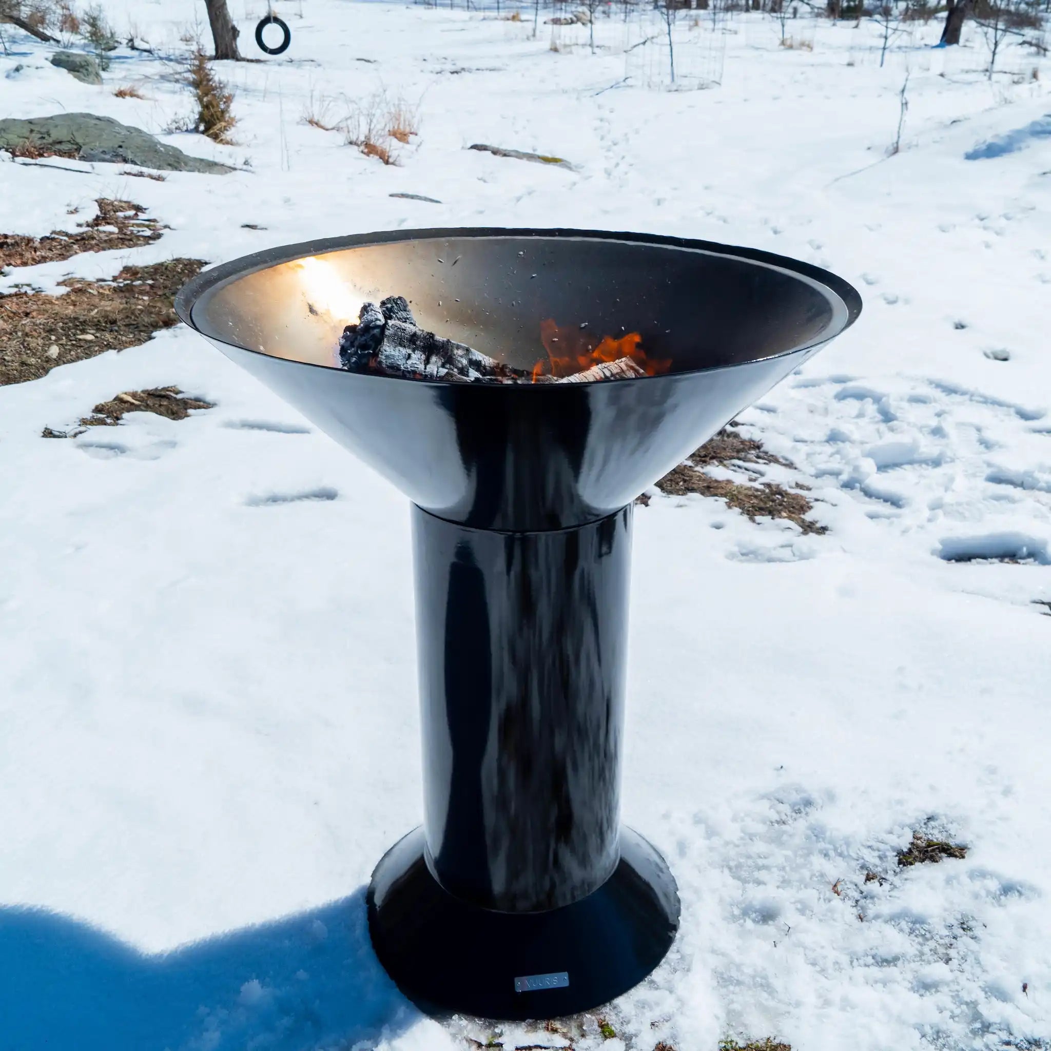 Vuuris Onyx outdoor fire pit in deep black porcelain enamel mounted on the large base with a fire inside against a background of a forest in the snow
