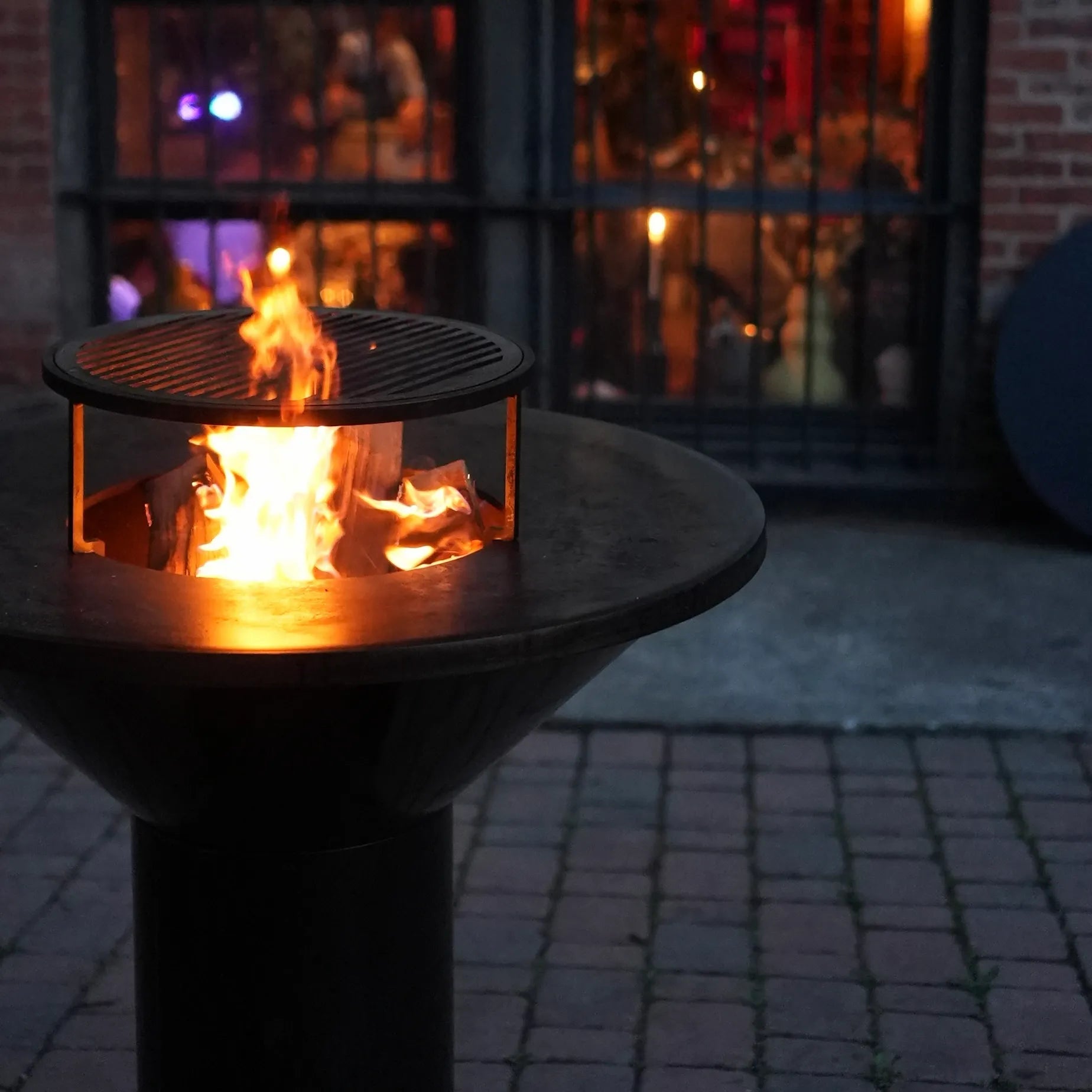 Nighttime picture of black Vuuris Onyx with fire inside and grill on top of cookingplate against a background of the window of a restaurant with people inside