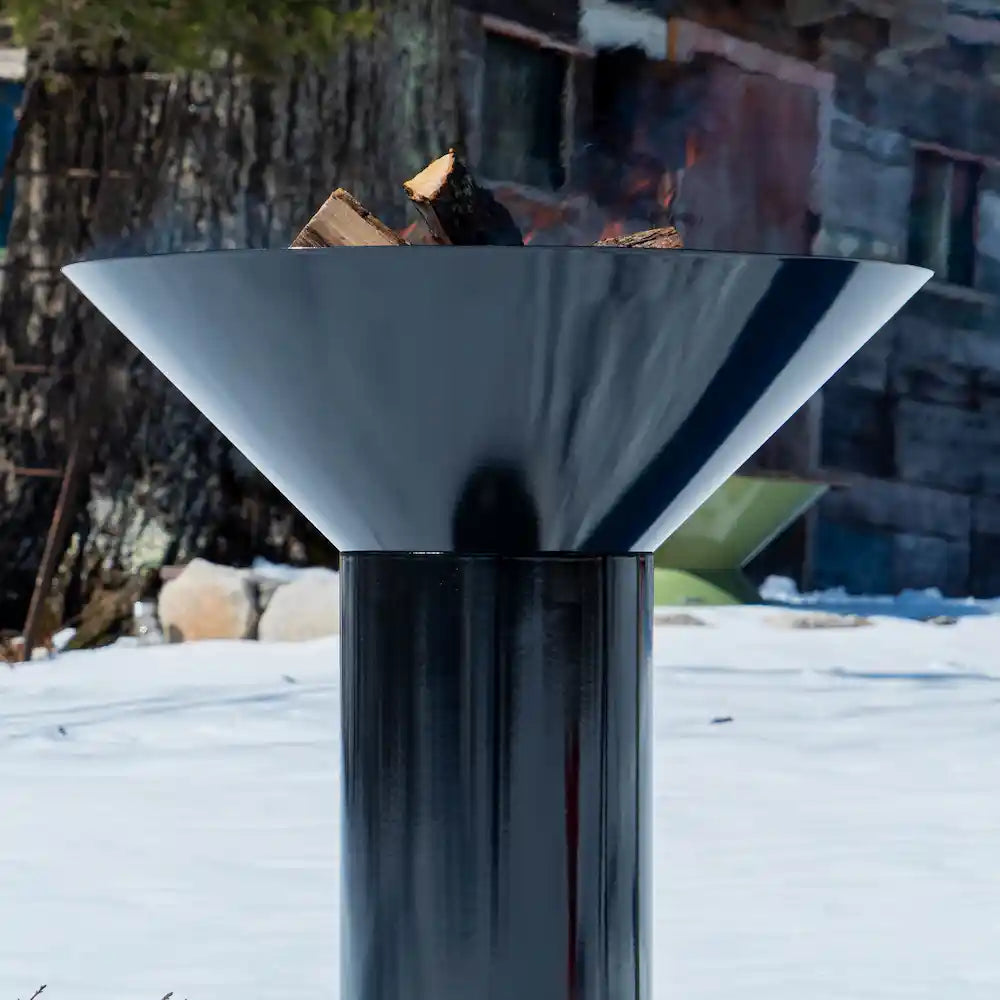 close up of Vuuris Onyx outdoor fire pit in deep black porcelain enamel mounted on the large base with a fire inside against a background of a wooden shed in the snow