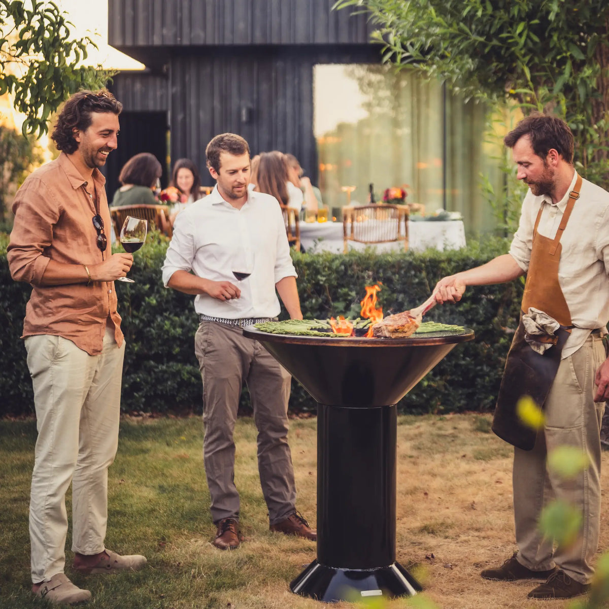 Three men standing outdoors around a black pedestal fire pit Onyx Vuuris with flames, in a backyard setting with a house and patio visible in the background.