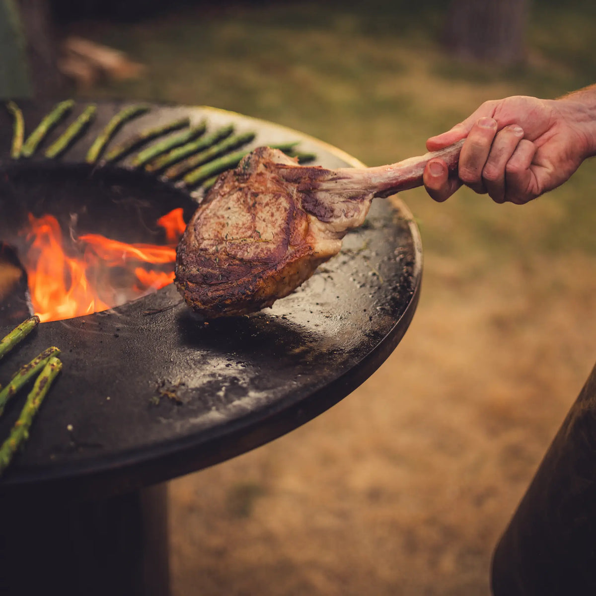 A hand holds a cooked bone-in steak over a black outdoor Vuuris Onyx grill with visible flames and asparagus, set in a real outdoor environment.