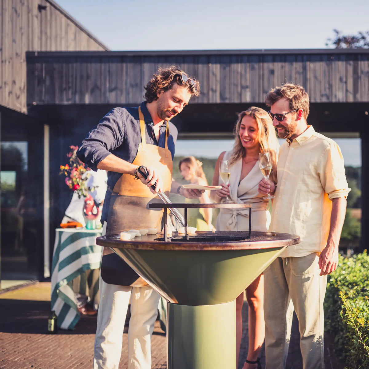 An outdoor lifestyle scene at a green Vuuris Sage barbecue grill with three people gathered around . A man on the left wears an apron and tends the grill, while two people on the right hold champagne glasses and smile.