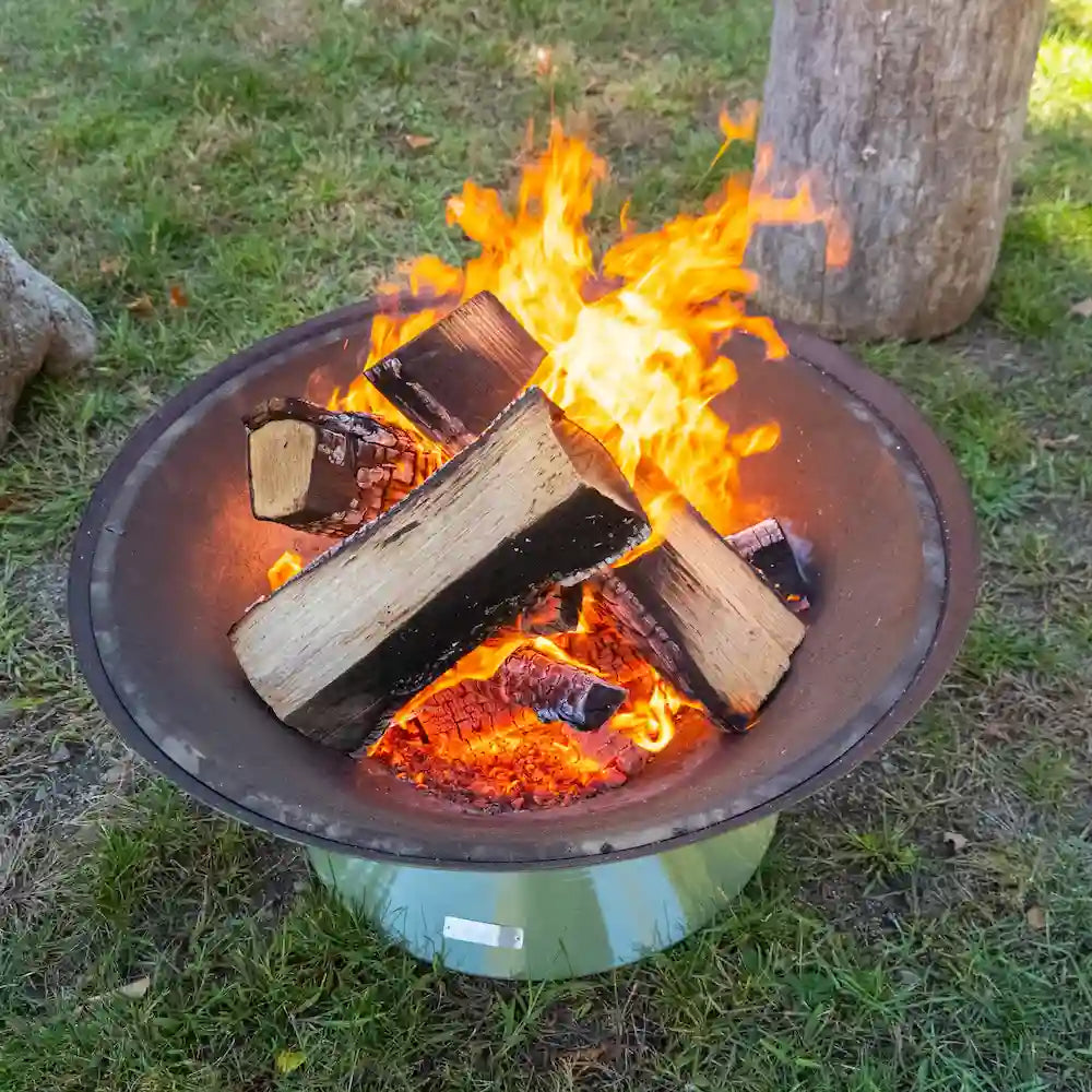 close up of green Vuuris Sage outdoor fire pit in earthy green porcelain enamel mounted on small base at lounge height with a fire inside, standing on grass in a garden