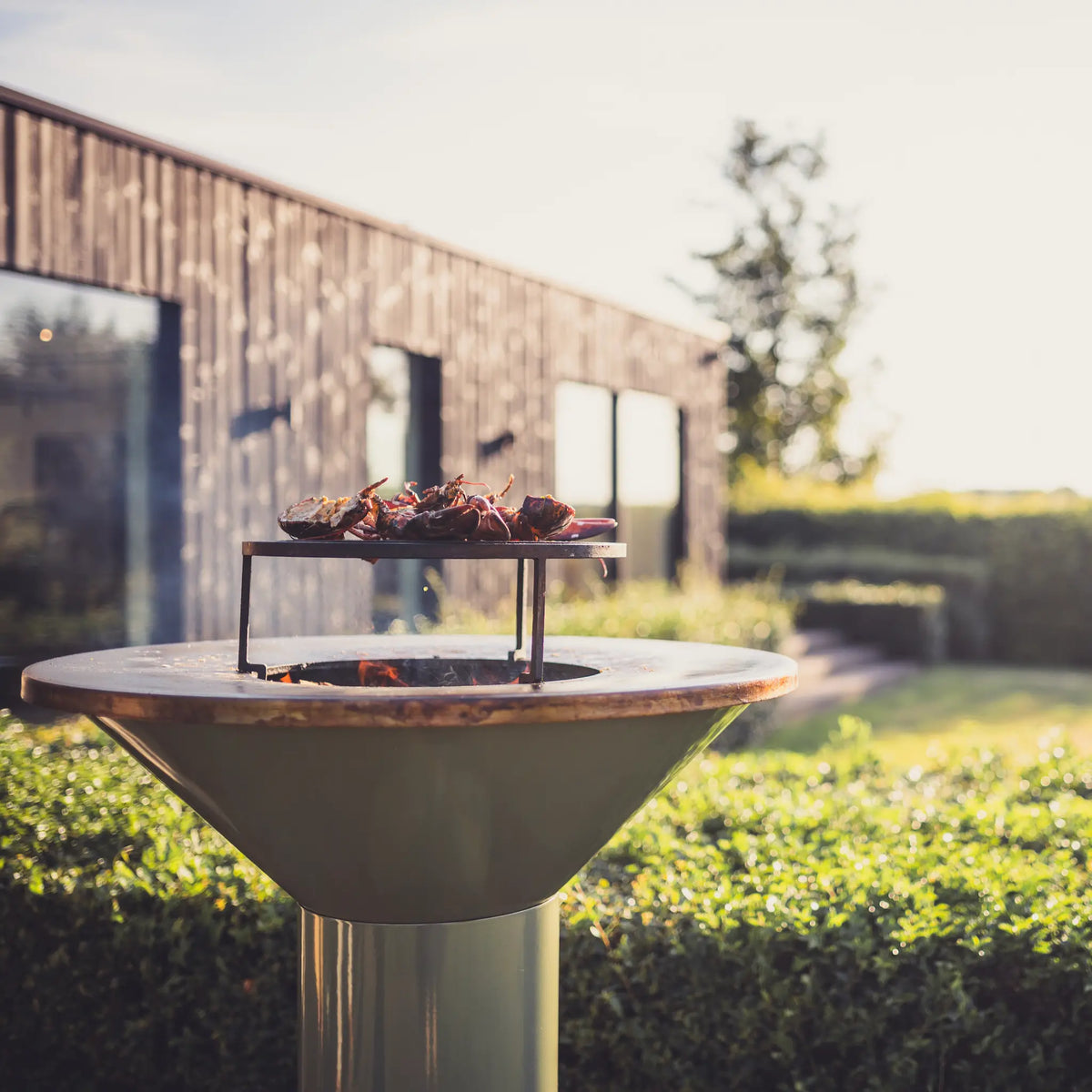 A modern outdoor patio Vuuris Sage fire pit bbq t with a wide enameled metal bowl and a small upper grill holding lobsters, set in a garden with a wooden house blurred in the background.