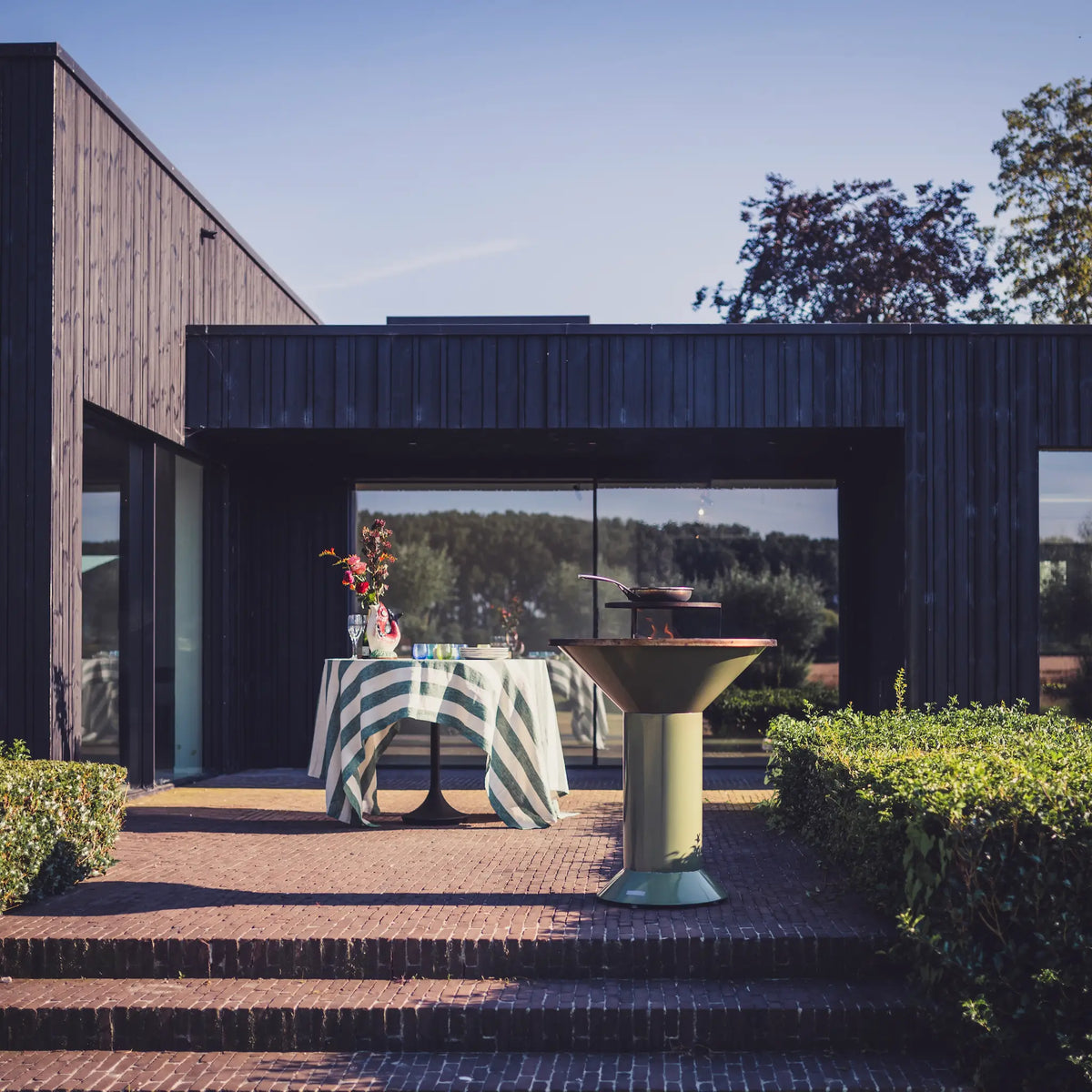 Outdoor dining setup on a brick patio in front of a modern dark wood building, featuring a striped tablecloth-covered table with flowers and a Vuuris Sage bbq in the foreground with grill and pan on top
