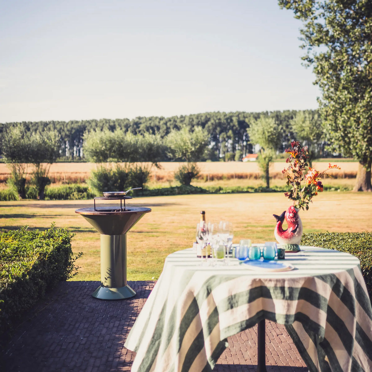An outdoor patio scene with a round table covered in a striped white tablecloth, set with glassware, a bottle, and a pink fish-shaped vase holding orange flowers. A metal pedestal Vuuris Sage bqq stands nearby, with a grassy field, trees, and a clear sky in the background.