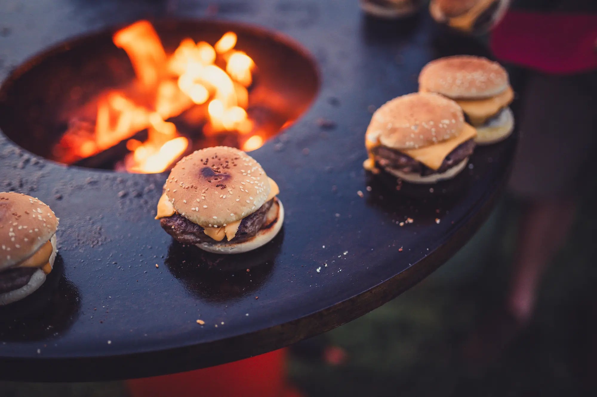 Small cheeseburgers cooking on a large black Vuuris Onyx outdoor grill with an open fire in the middle of the cooking plate.