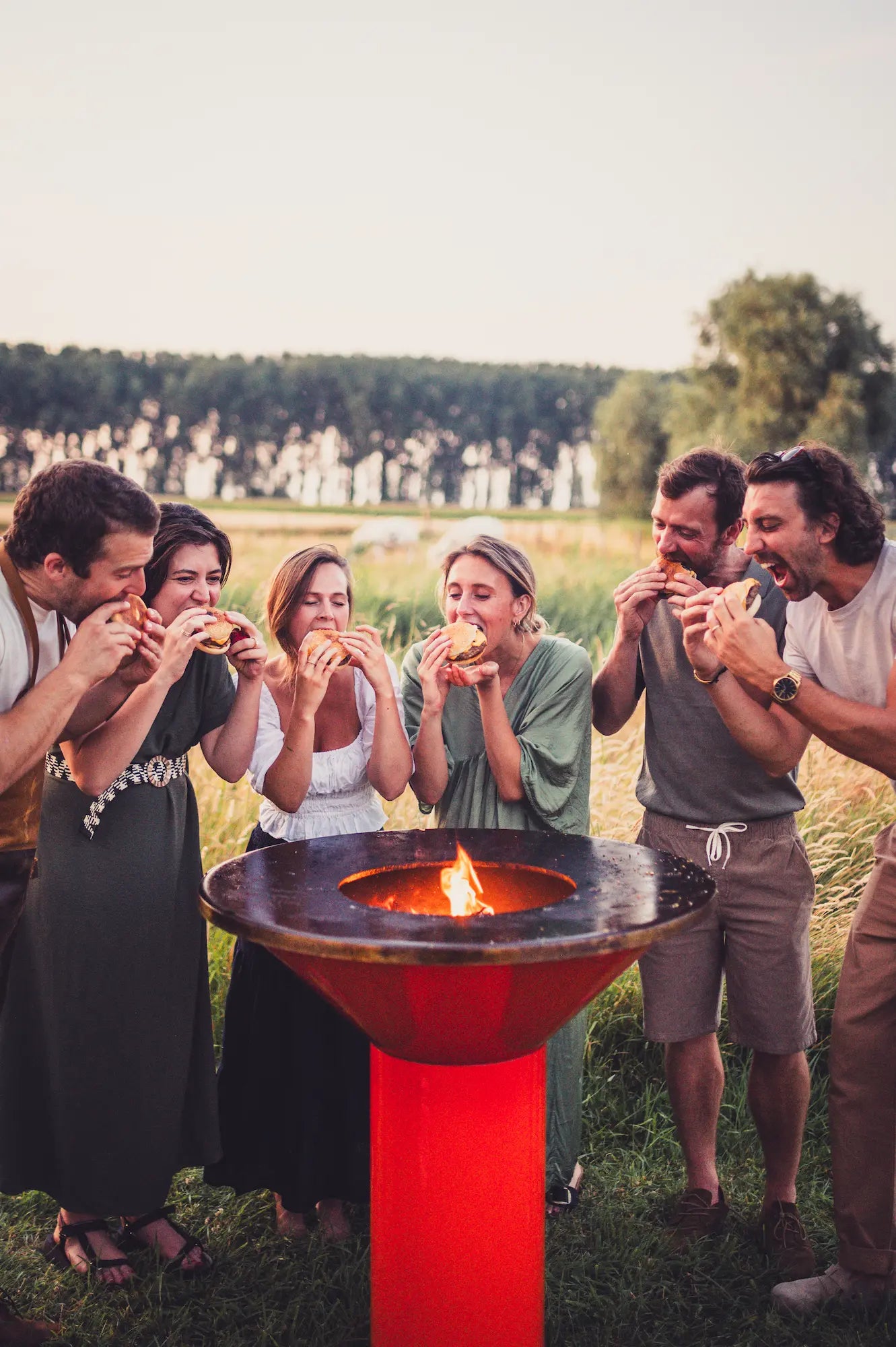 A group of six friends gathered outdoors in a grassy field, sharing a burger while standing around a modern orange Vuuris Tiger with black cooking plate and fire with a visible flame in the middle