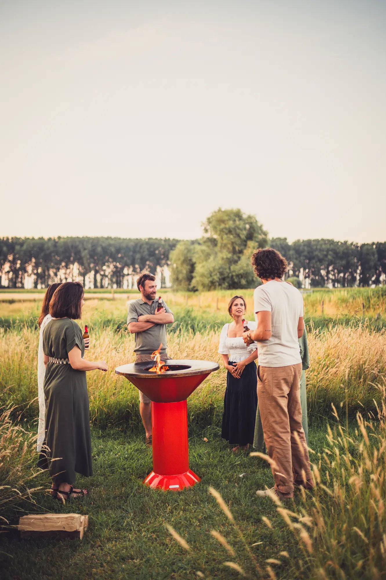 Four people standing outdoors in a grassy field around a Vuuris Tiger fire pit with flames burning inside. The scene is shot in warm golden-hour light with trees in the background.
