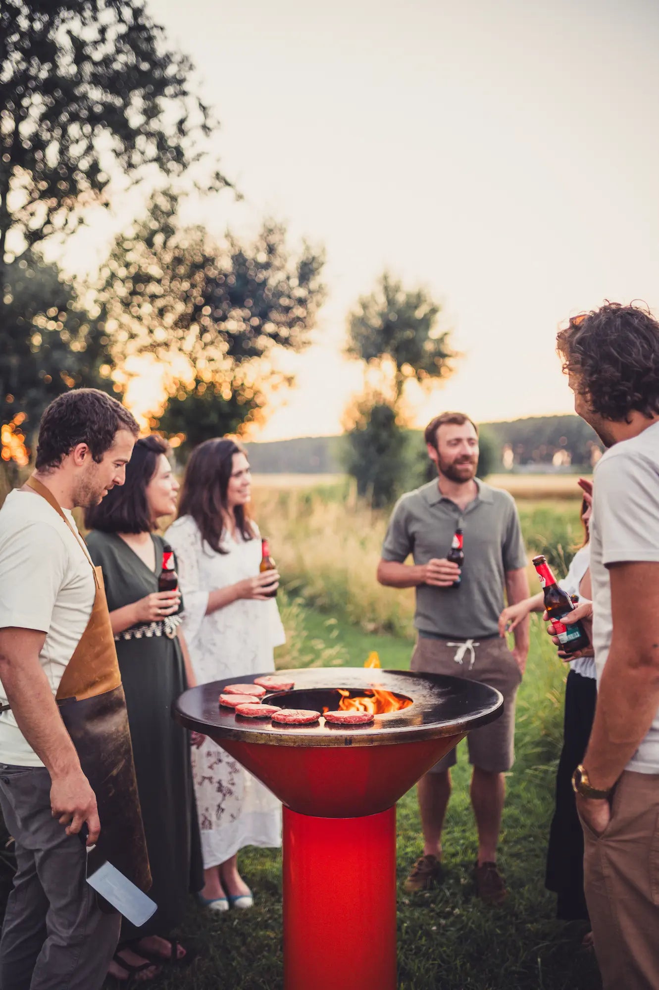 An outdoor social scene at sunset with several people gathered around a freestanding orange Vuuris Tiger round barbecue grill with food cooking on top, holding drinks in a grassy field.