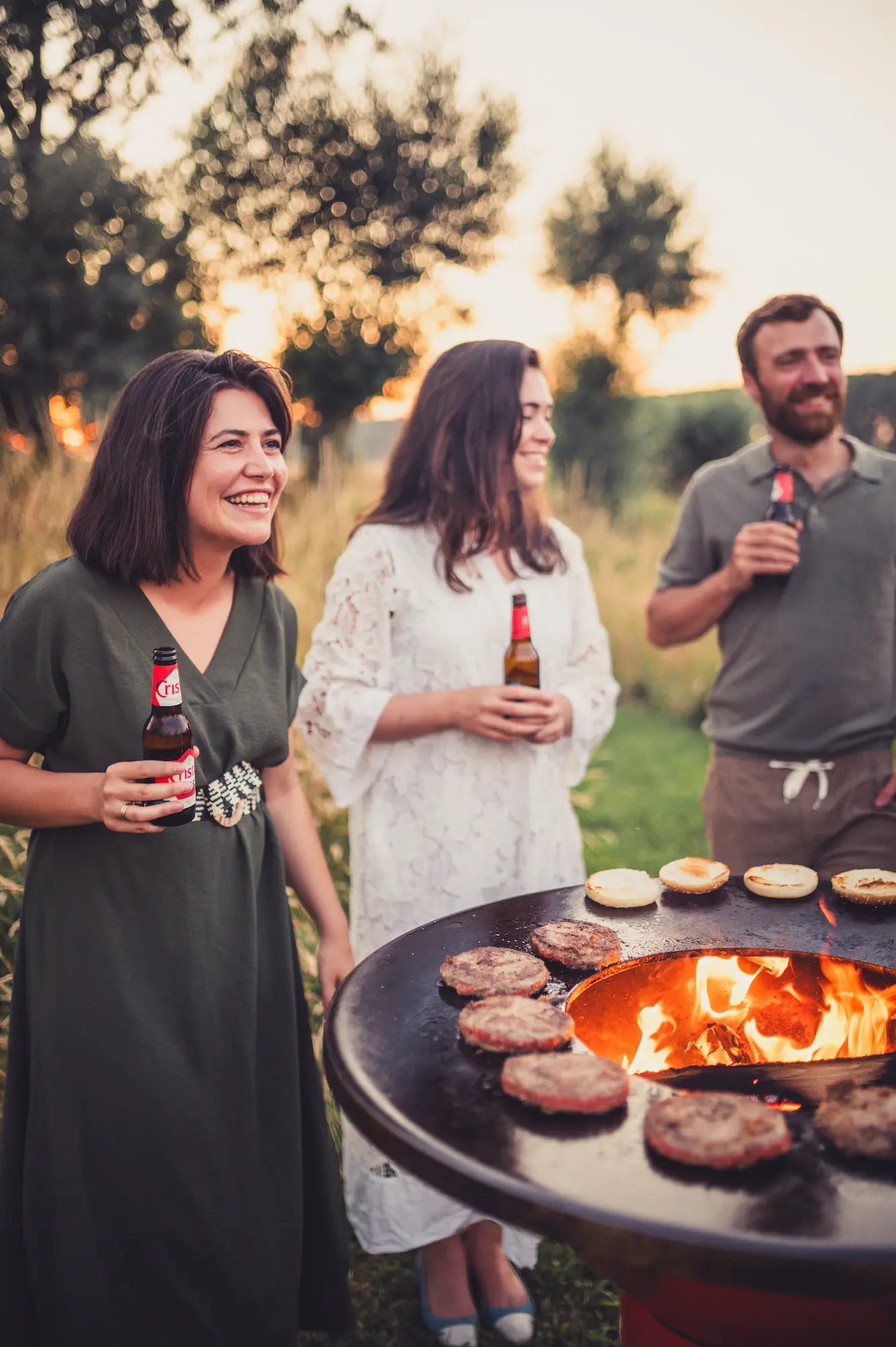 Three people standing outdoors by a round Vuuris Tiger fire pit grill with burgers cooking, each holding a beer bottle, set against a warm sunset background.