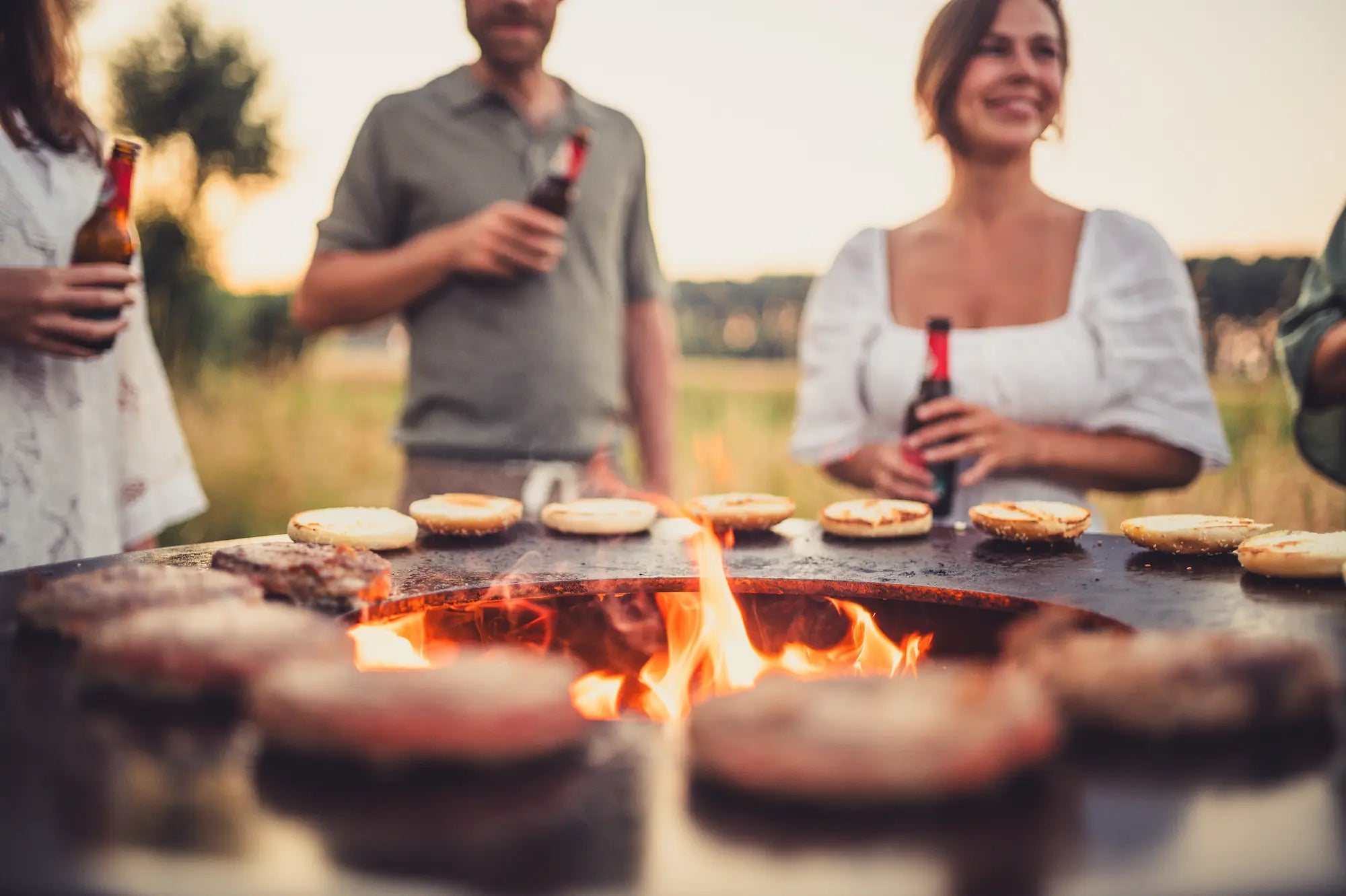 Outdoor barbecue scene at sunset with people holding beer bottles behind a round Vuuris Tiger grill where burgers and buns are cooking over visible flames.