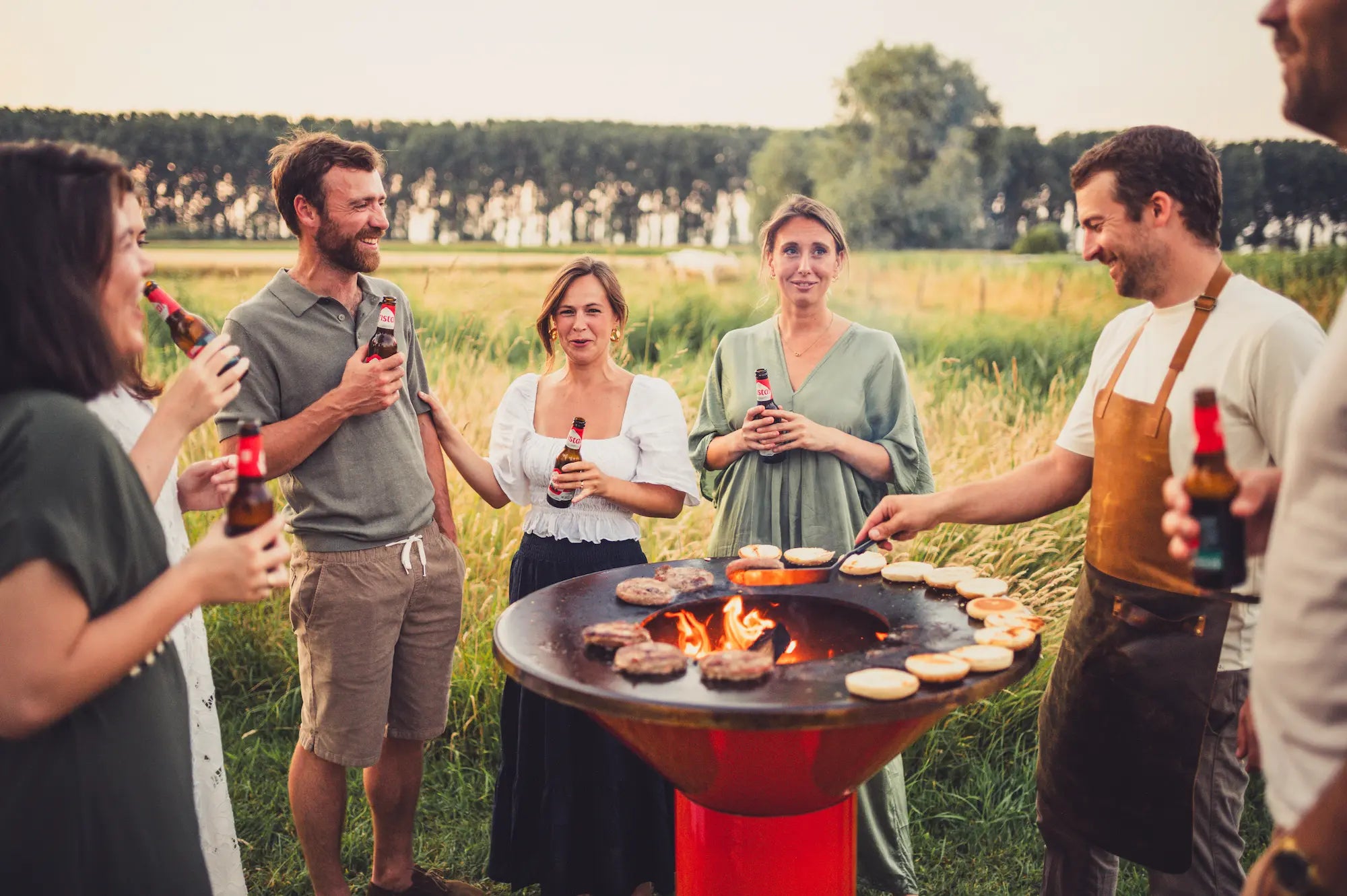 A group of friends gathered outdoors around an orange Vuuris Tiger barbecue grill, cooking burgers and flipping buns over an open flame in a grassy field at sunset.