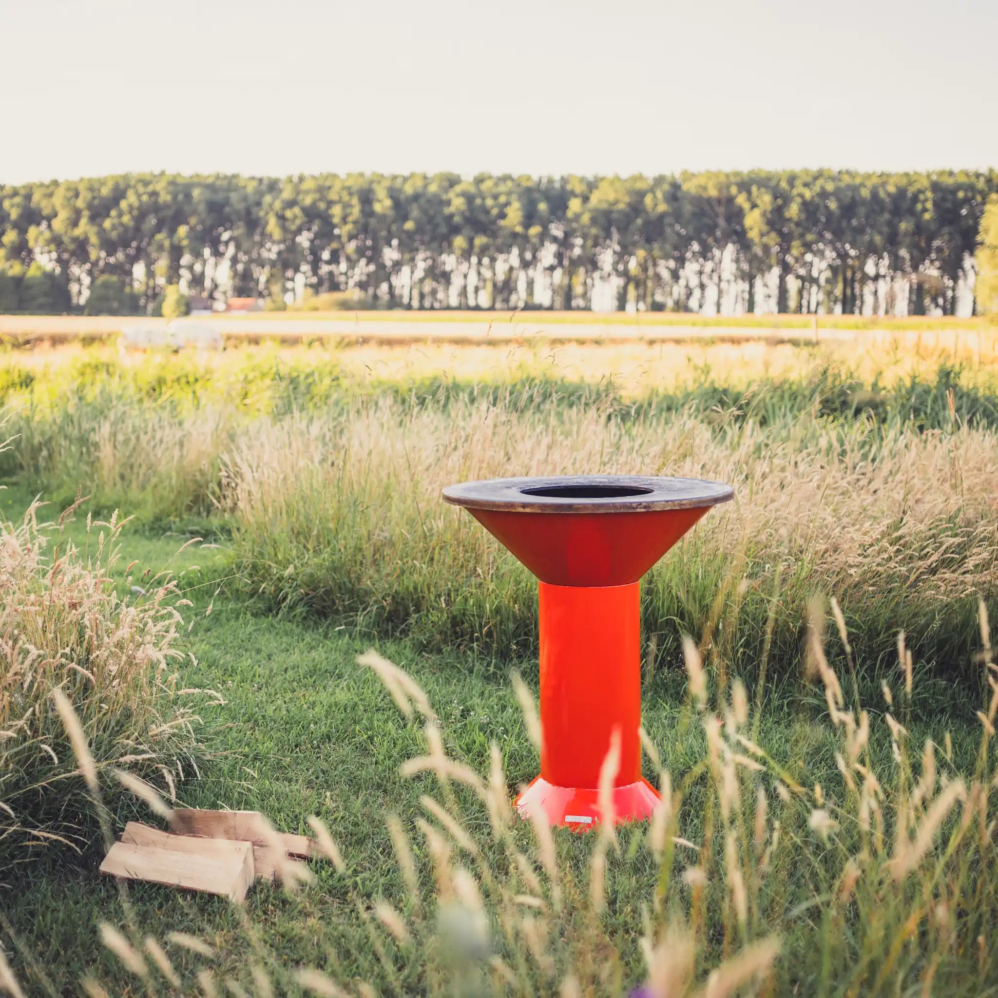 An orange Vuuris Tiger outdoor bbq, set in a grassy field with tall dry grasses and a line of trees in the background.