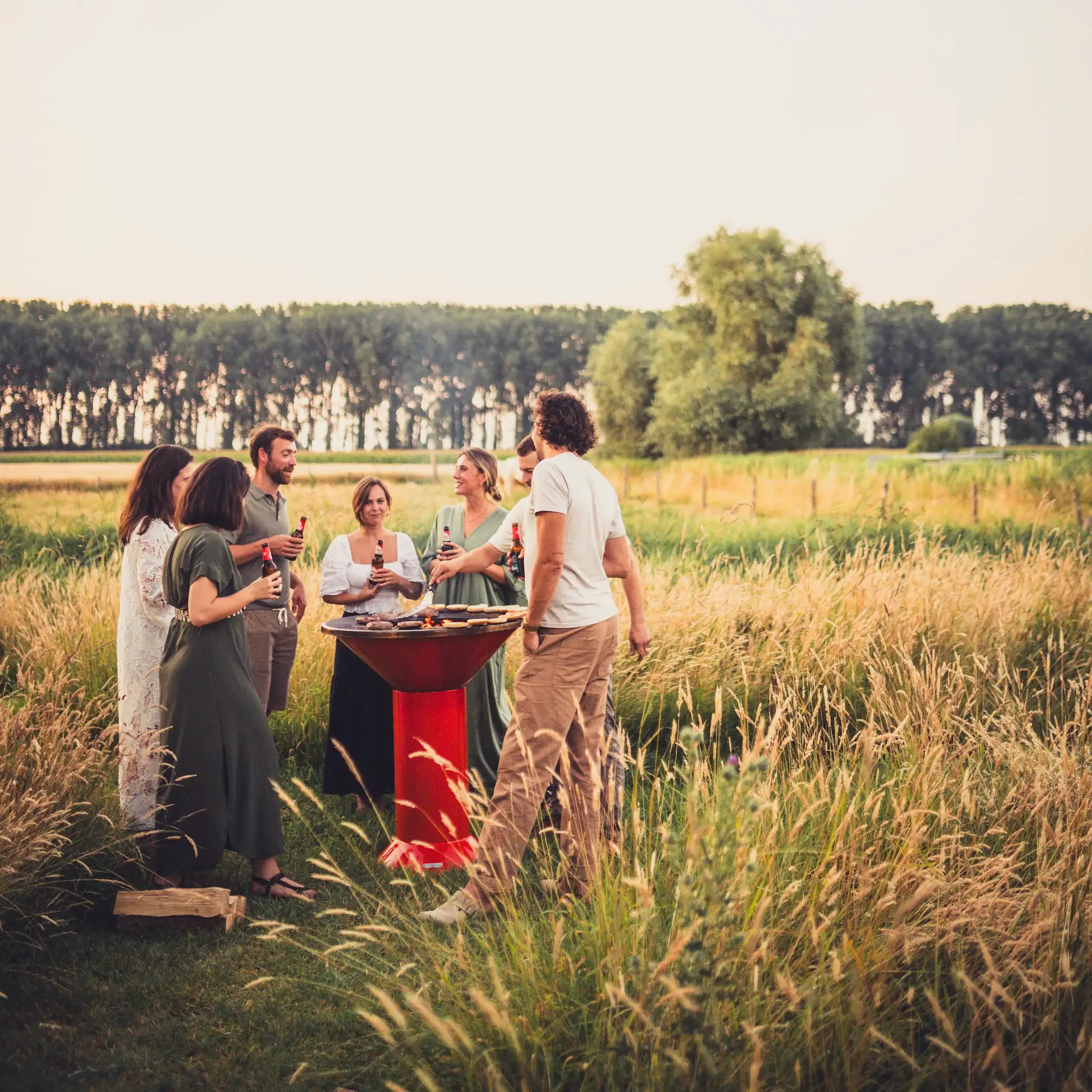 A group of people gathered outdoors in a grassy field near an orange Vuuris Tiger bbq fire pit grill with trees in the background under a pale sky.