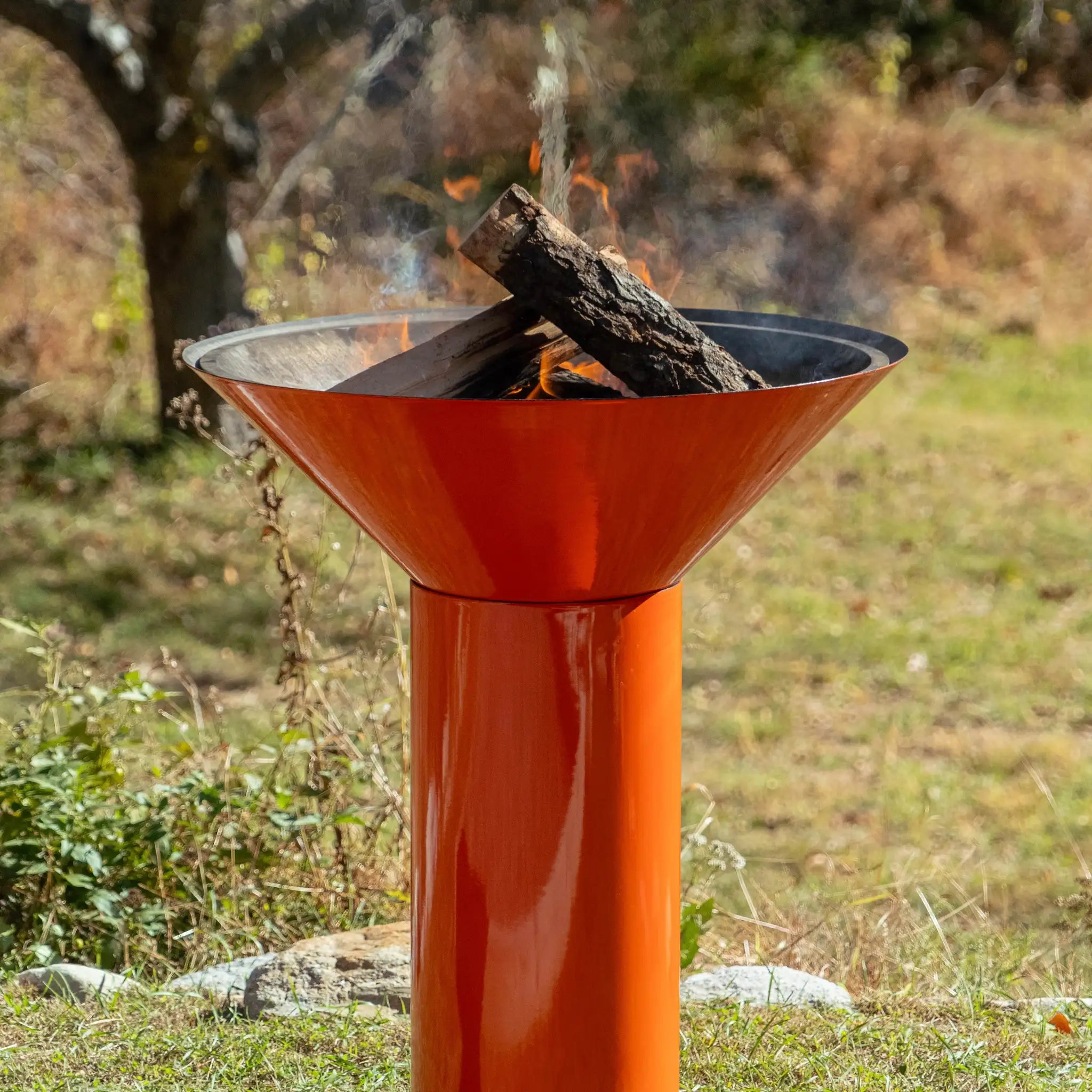 Vuuris Tiger outdoor fire pit in vibrant orange porcelain enamel on large base at counterheight against a background of a grassy garden in the summer. there is a fire burning in the pit