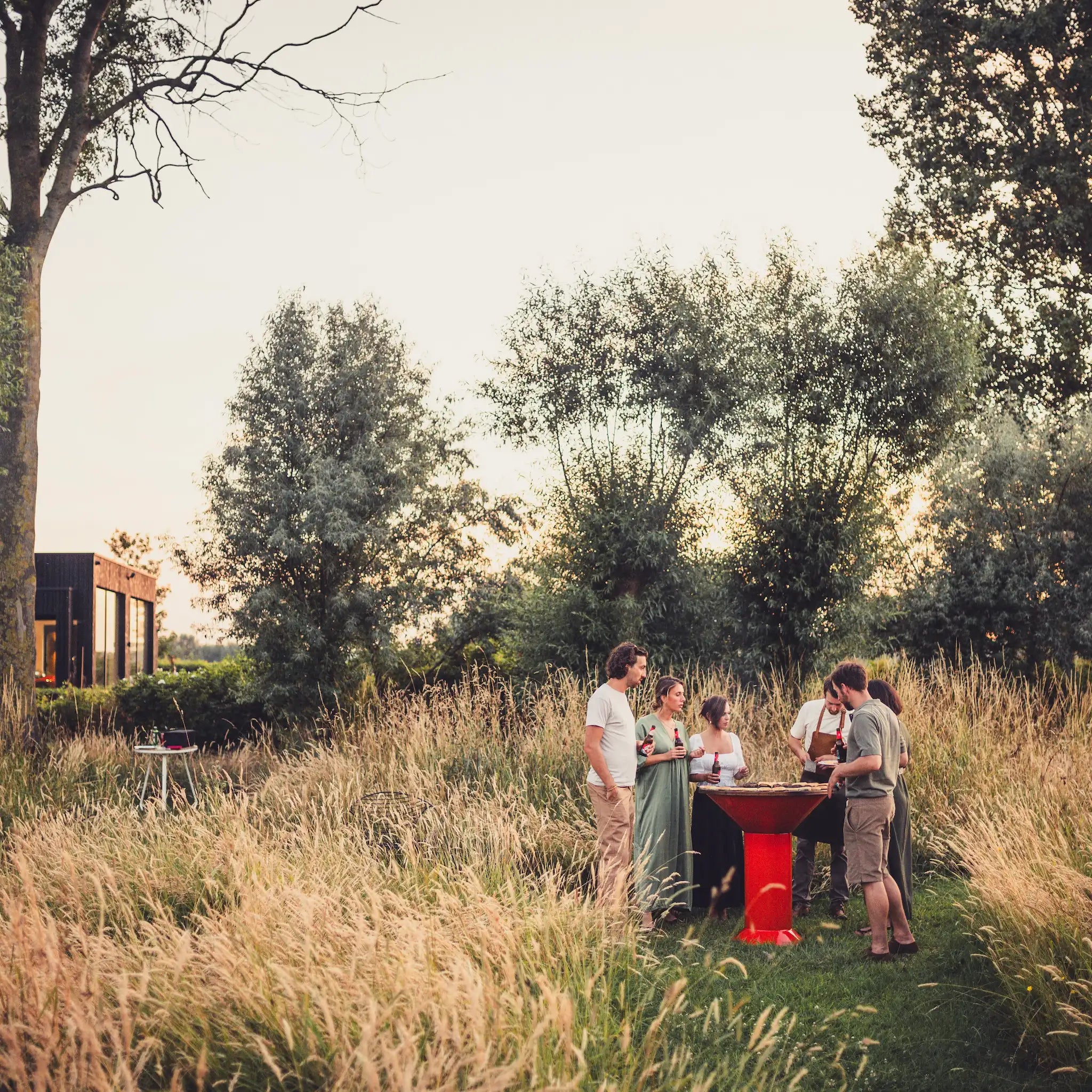 An outdoor lifestyle scene in a grassy field at sunset, showing a small group of people gathered around an orange Vuuris Tiger bbq near trees and a modern house in the background.