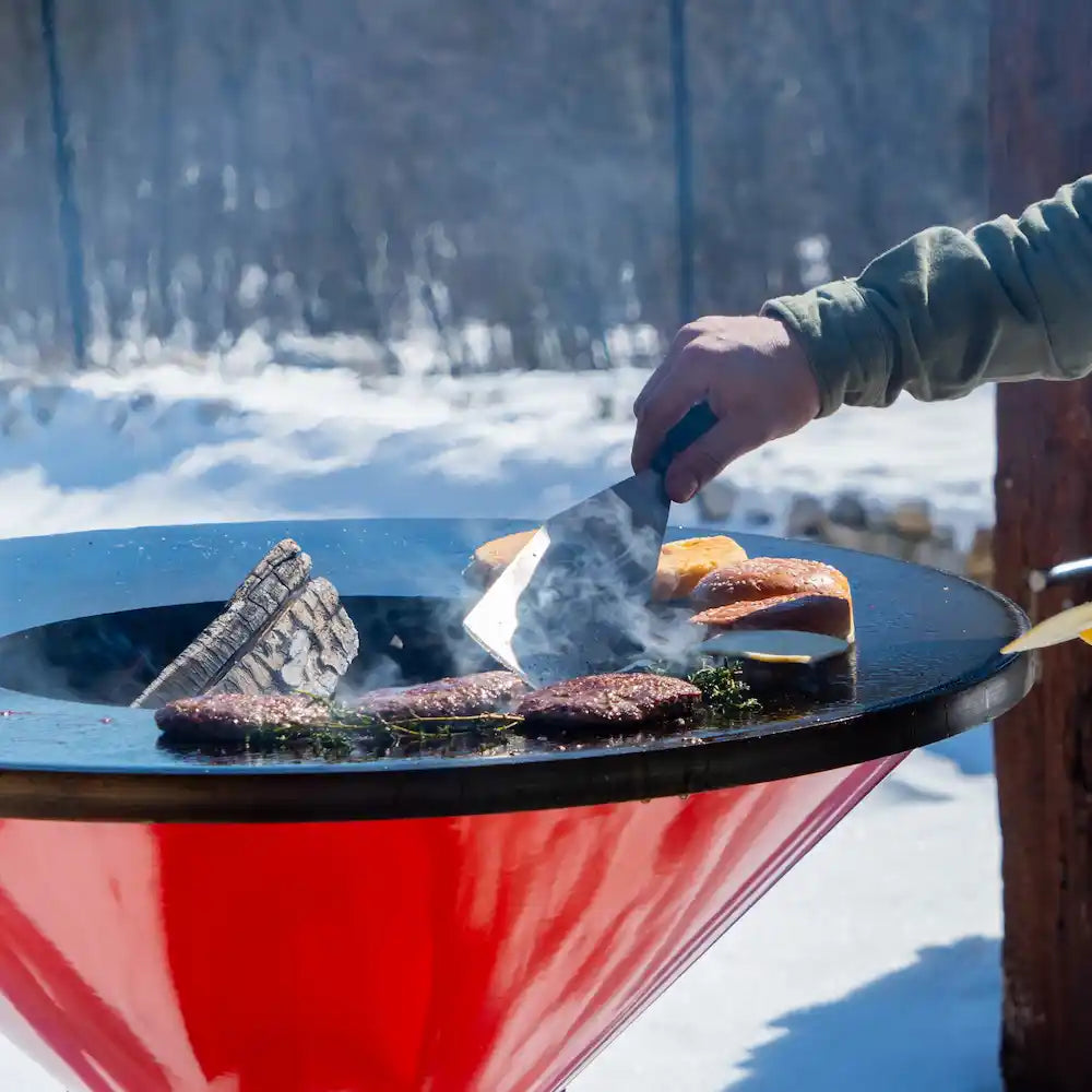 Person grilling burgers on an orange Vuuris on the cooking plate with a snowy landscape in the background